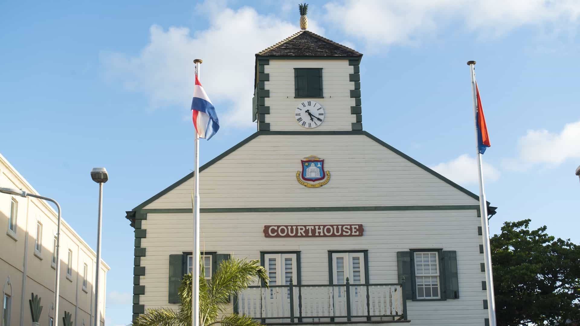 A photograph of the historic Sint Maarten Courthouse in Philipsburg, St. Maarten. The striking white colonial-style building features a prominent, circular structure topped by a small, wooden cupola with a pineapple finial (a traditional symbol of hospitality). The courthouse stands directly on the street, near the waterfront, against a blue sky.