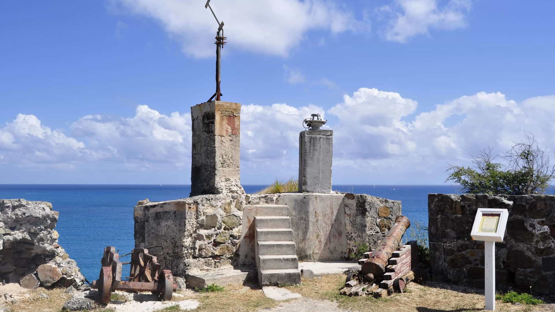 A scenic view of the ruins of Fort Amsterdam in St. Maarten, situated on a peninsula overlooking the Caribbean Sea. The image shows remnants of stone walls and aged cannons pointing towards the turquoise water. The fort's location offers a panoramic view of the harbor and the nearby beaches.c