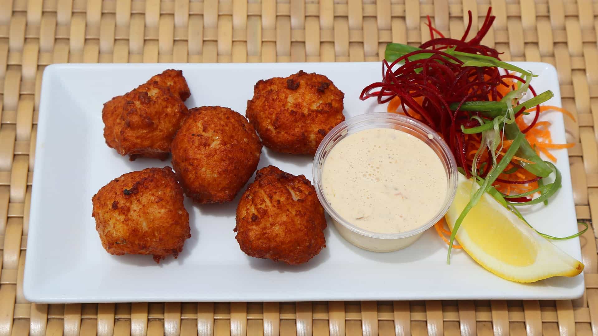 A close-up, appetizing photograph of a plate of deep-fried Conch Fritters, a popular seafood snack. The golden-brown, irregularly shaped fritters are piled high and served with a small bowl of pink or orange dipping sauce (likely a spicy aioli or chili sauce). A garnish of fresh parsley or cilantro is scattered over the fritters.
