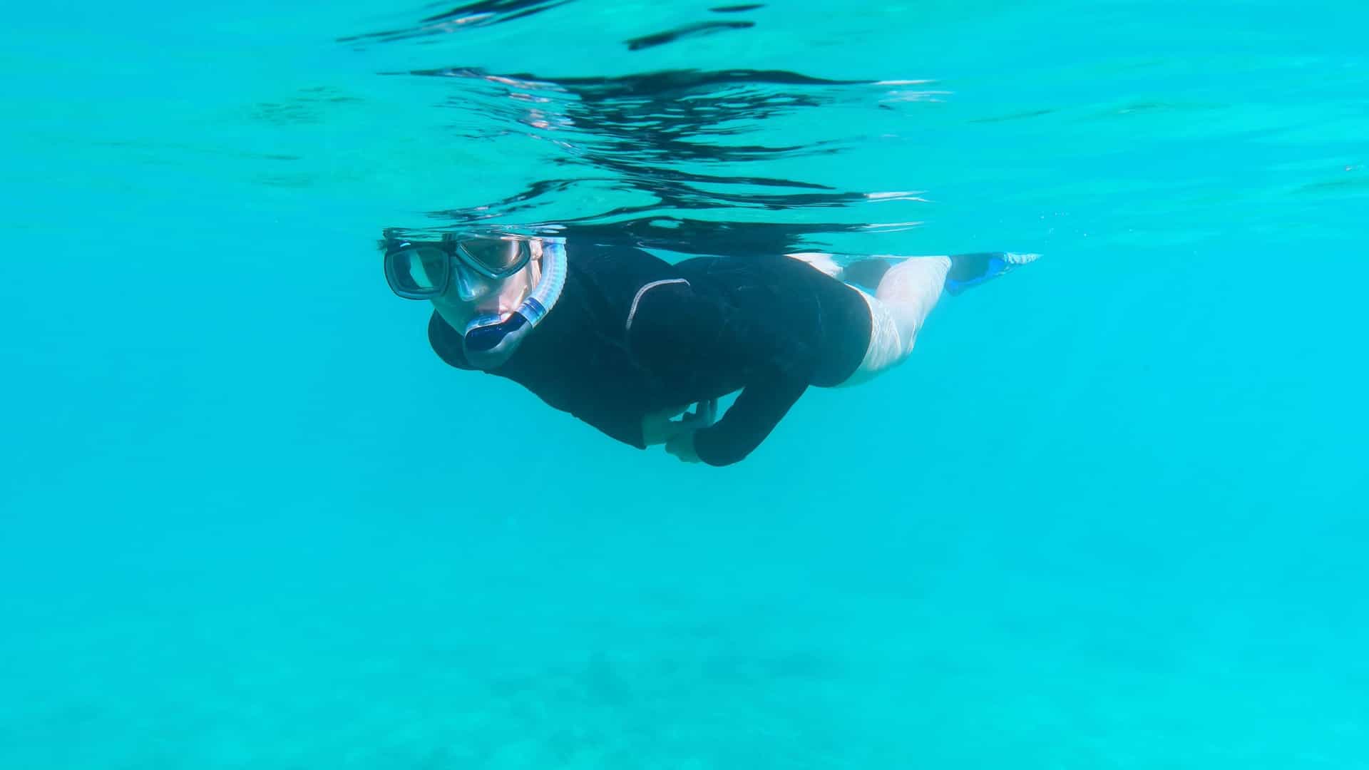 A bright, underwater view showing a person enjoying a snorkel in the crystal-clear waters of the Caribbean. The surface of the water, illuminated by strong sunlight, is visible above the snorkeler. Below, the seabed is covered with a vibrant coral reef and rocky outcroppings, with small tropical fish swimming near the coral.
