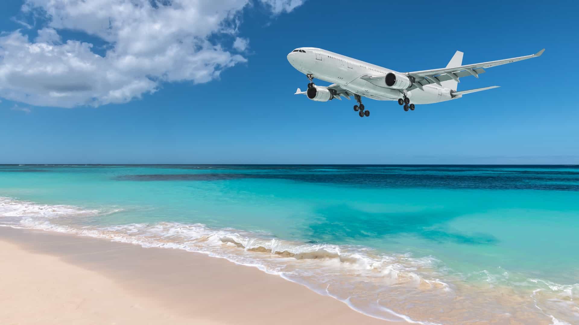 A large commercial airplane descends for a low altitude landing over Maho Bay Beach in St. Maarten. The scene, viewed from the beach, shows the plane flying directly over beachgoers, with the vibrant turquoise water and white sand of the Caribbean beach visible below.