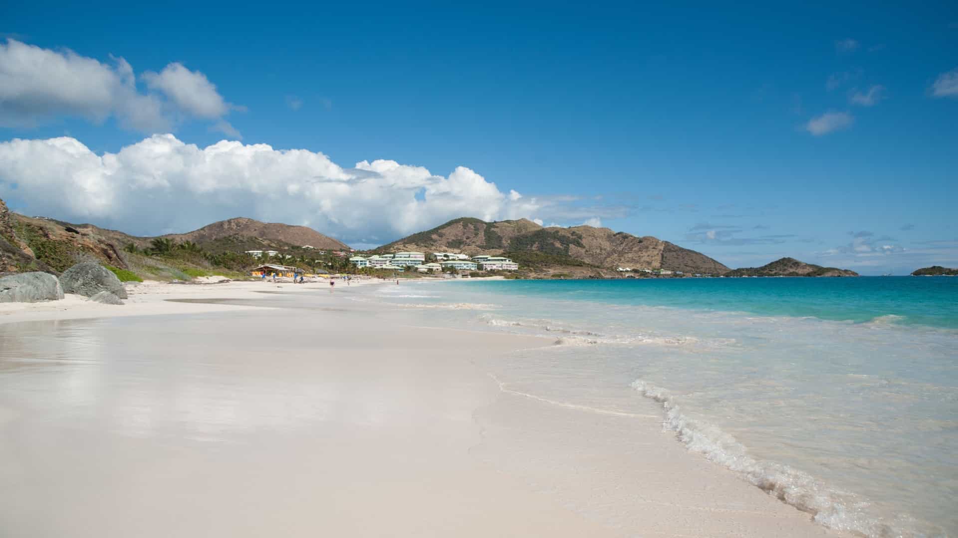 A pristine, panoramic view of Orient Bay Beach on the French side of Saint Martin. The image captures a wide expanse of fine white sand curving along a turquoise bay. The calm, clear water is a vibrant blue, and the beach is lined with palm trees and green hills in the background.
