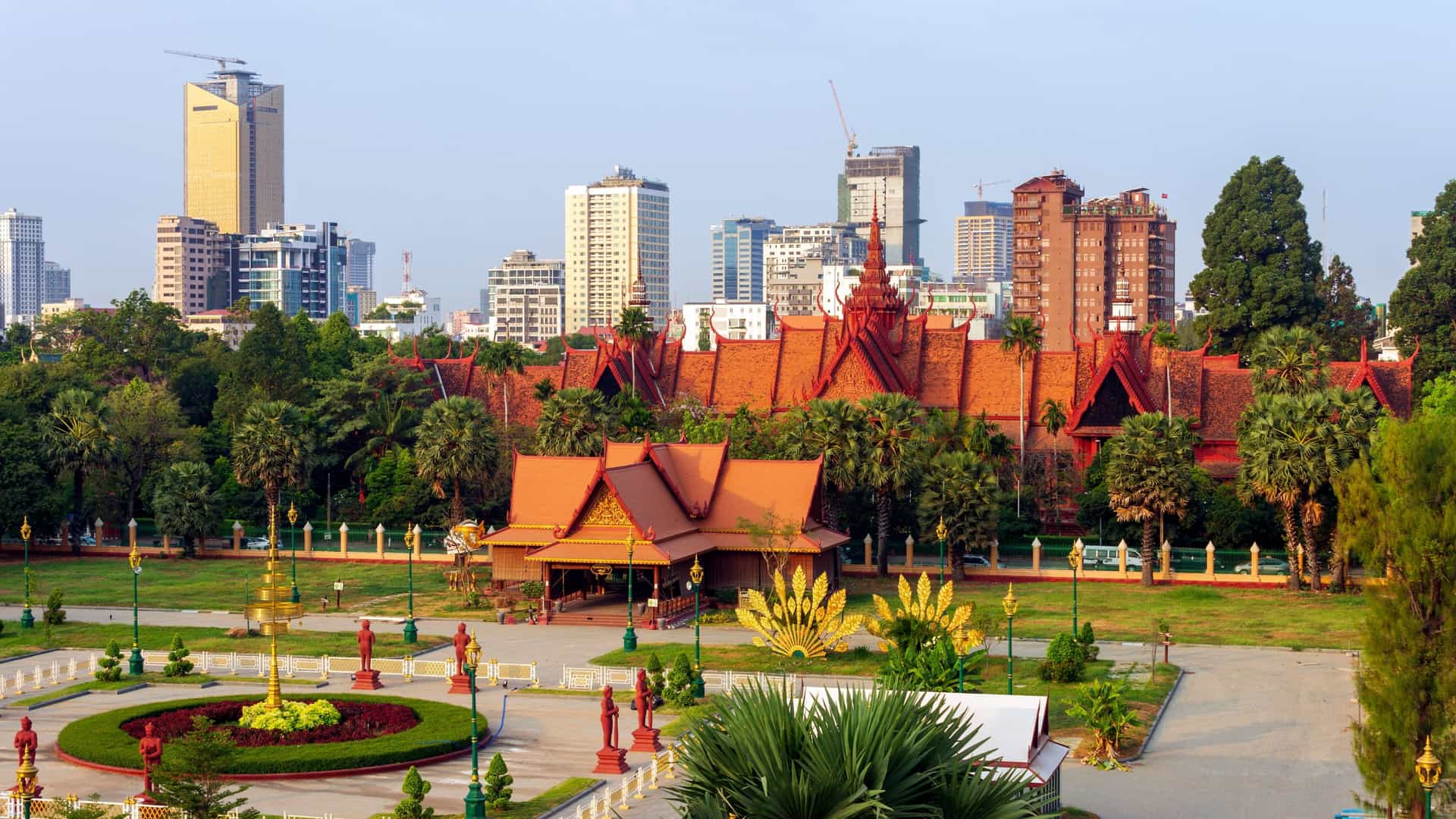 The National Museum of Cambodia in Phnom Penh, a building constructed in a traditional Khmer architectural style with red sandstone and a tiered, tiled roof. The facade features elaborate carvings and a central tower, and it is framed by a lush, green courtyard and manicured plants.