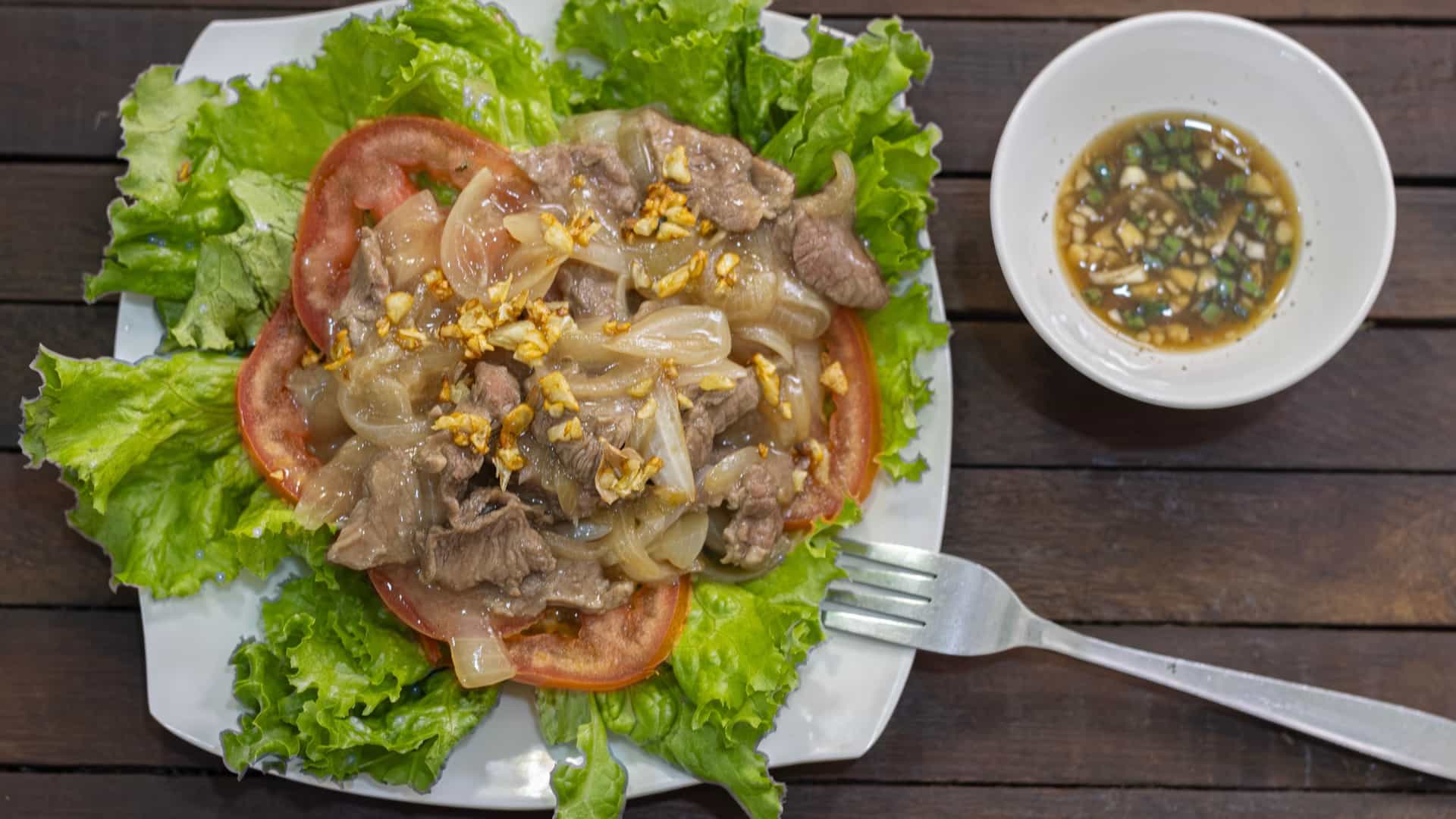 A close up, top down view of a plate of traditional Cambodian Lok Lak, or "shaking beef." The dish consists of cubes of glossy, stir fried beef and sliced onions, served on a bed of crisp green lettuce and sliced red tomatoes. A small dish of dipping sauce, made from lime juice, salt, and pepper, is placed on the side. The dish is presented on a modern white plate.