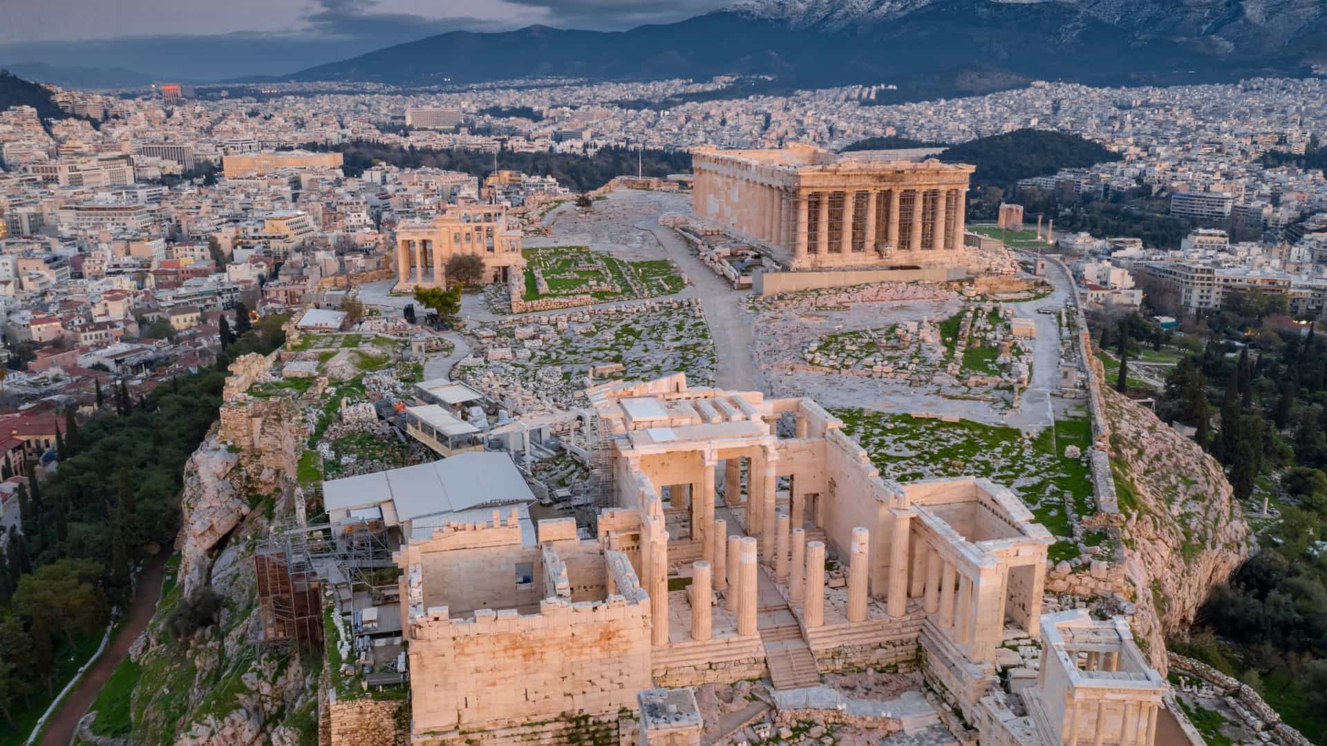 An aerial view of the Acropolis of Athens at sunset. The ancient citadel is a rocky outcrop rising above the city, with several iconic structures visible on its summit, including the Temple of Athena Nike, the Parthenon, and the Erechtheion. The city of Athens spreads out in the background under the golden light of the setting sun.
