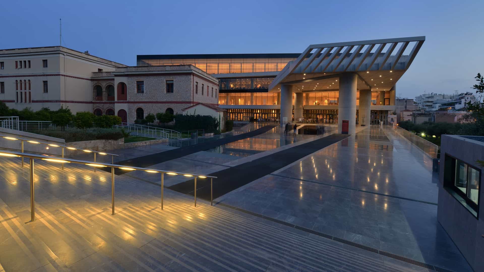 A night view of the Acropolis Museum in Athens, Greece. The modern building, with its striking glass and steel facade, is illuminated against the dark sky. The lights from inside highlight the architectural design, and the urban landscape of Athens is visible in the background.
