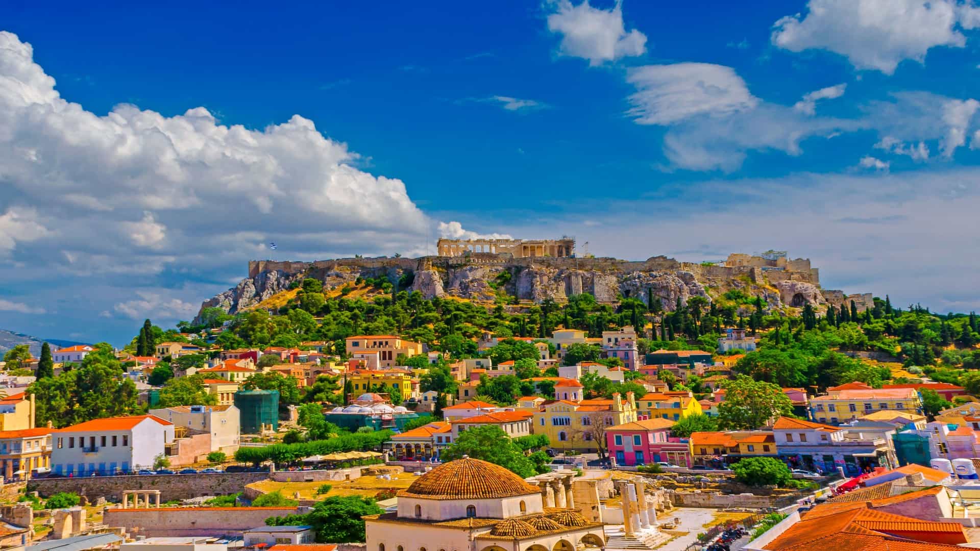 An aerial view of the historic Plaka neighborhood in Athens, Greece, with the Acropolis prominently featured on its rocky hill. The dense residential buildings of Plaka, characterized by their traditional architecture, fill the foreground and midground, leading up to the large, rocky outcrop of the Acropolis.
