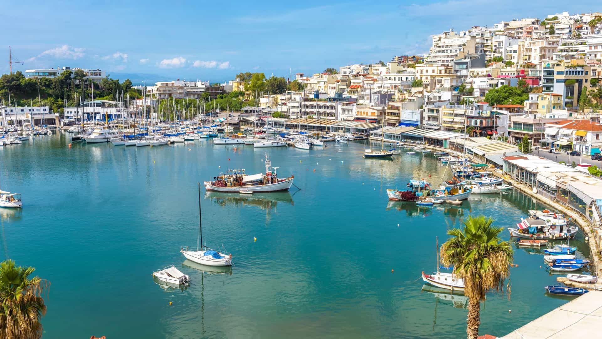 A scenic view of the Mikrolimano marina in Piraeus, Athens, Greece. Numerous small boats and sailboats are moored in the calm waters of the horseshoe shaped harbor, surrounded by buildings on the hillside.