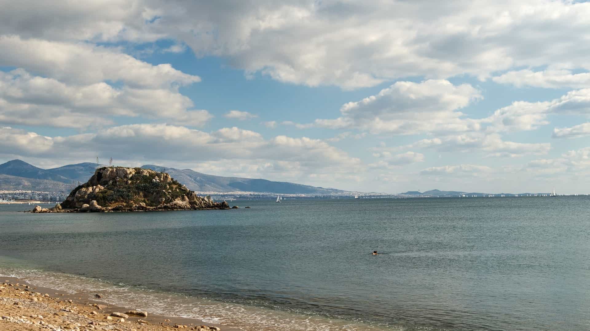A calm, scenic view of Votsalakia Beach in Piraeus, Greece, with no people on the shore. The beach, with its rocky and coarse sand, meets a tranquil body of water. In the background, a small, lushly vegetated island is visible against the horizon.
