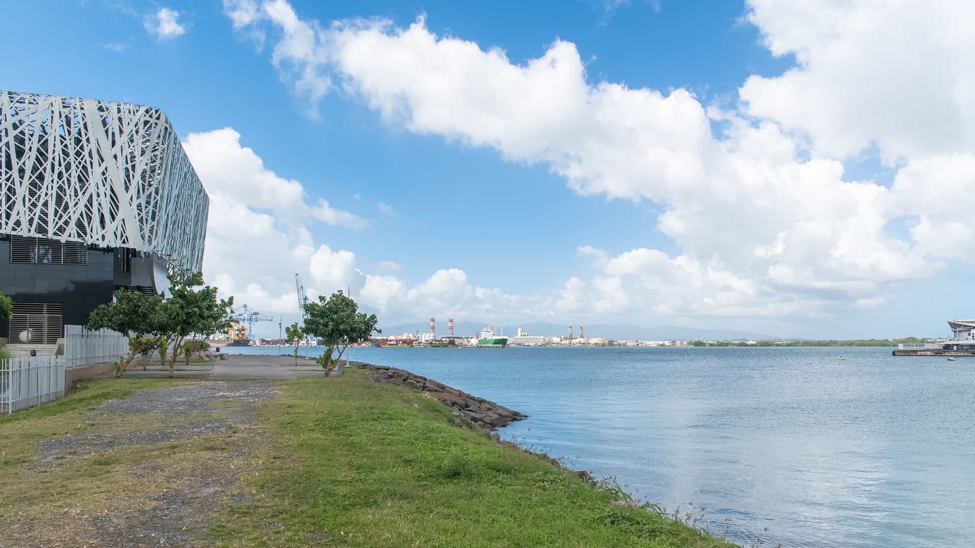 A scenic photograph of the waterfront in Pointe-à-Pitre, Guadeloupe. On the left is the distinctive modern architecture of the Mémorial ACTe, a large building encased in a black and white geometric lattice pattern. In the foreground, a grassy embankment meets the dark water of the harbor, while the industrial port and mountains are visible across the bay under a bright blue sky with large white clouds.