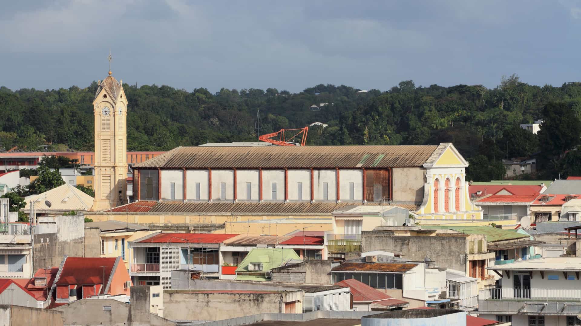A street-level photograph of the historic Cathedral of St. Pierre and St. Paul in Pointe-à-Pitre, Guadeloupe. The church is notable for its unique, dark green, open ironwork structure and yellow facade, earning it the nickname "Iron Cathedral." The ornate bell tower stands to the left of the main entrance, with surrounding palm trees and colonial buildings visible.