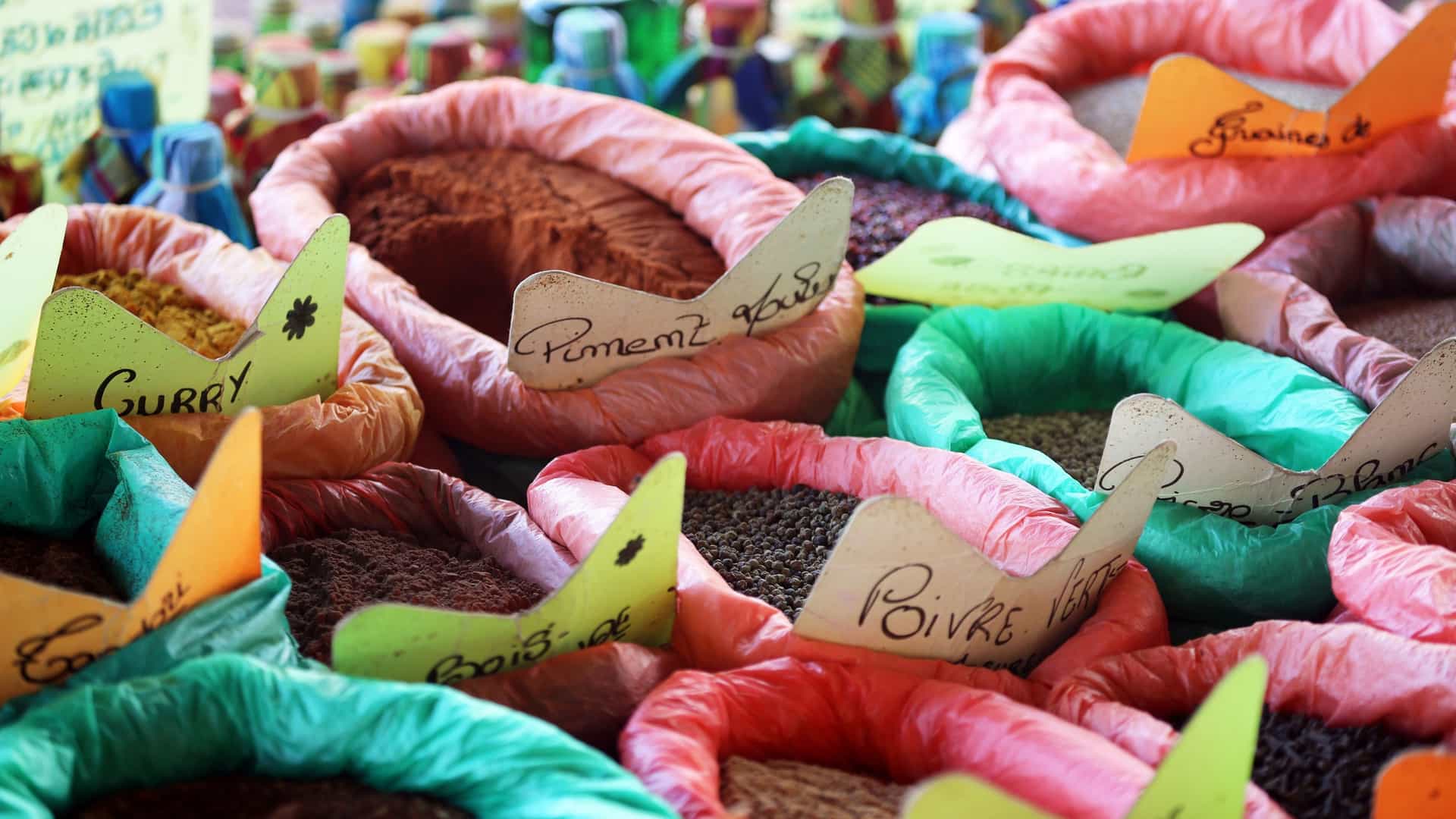 A close-up photograph of a vibrant Guadeloupe Spice Market stall displaying large, open bags filled with various whole and ground spices. The spices, including curry powder, peppercorns (Poivre), and allspice (Piment), are contained in bags made of brightly colored pink, red, and green plastic. Hand-written paper labels identify the different spices in French.