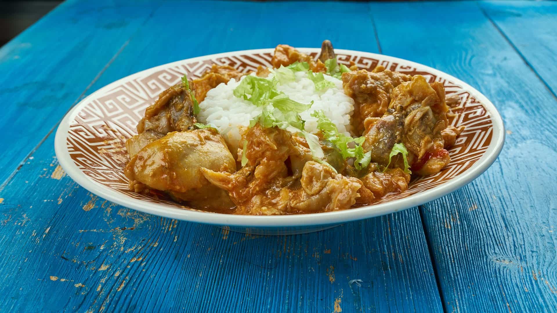 A close-up view of a bowl of Colombo de Poulet, a vibrant, stewed chicken dish from the Caribbean. The curry-like dish features chunks of chicken, potatoes, and other vegetables in a rich, yellowish-orange sauce made with colombo powder (a spice blend). The bowl of chicken is served alongside a separate portion of white rice.