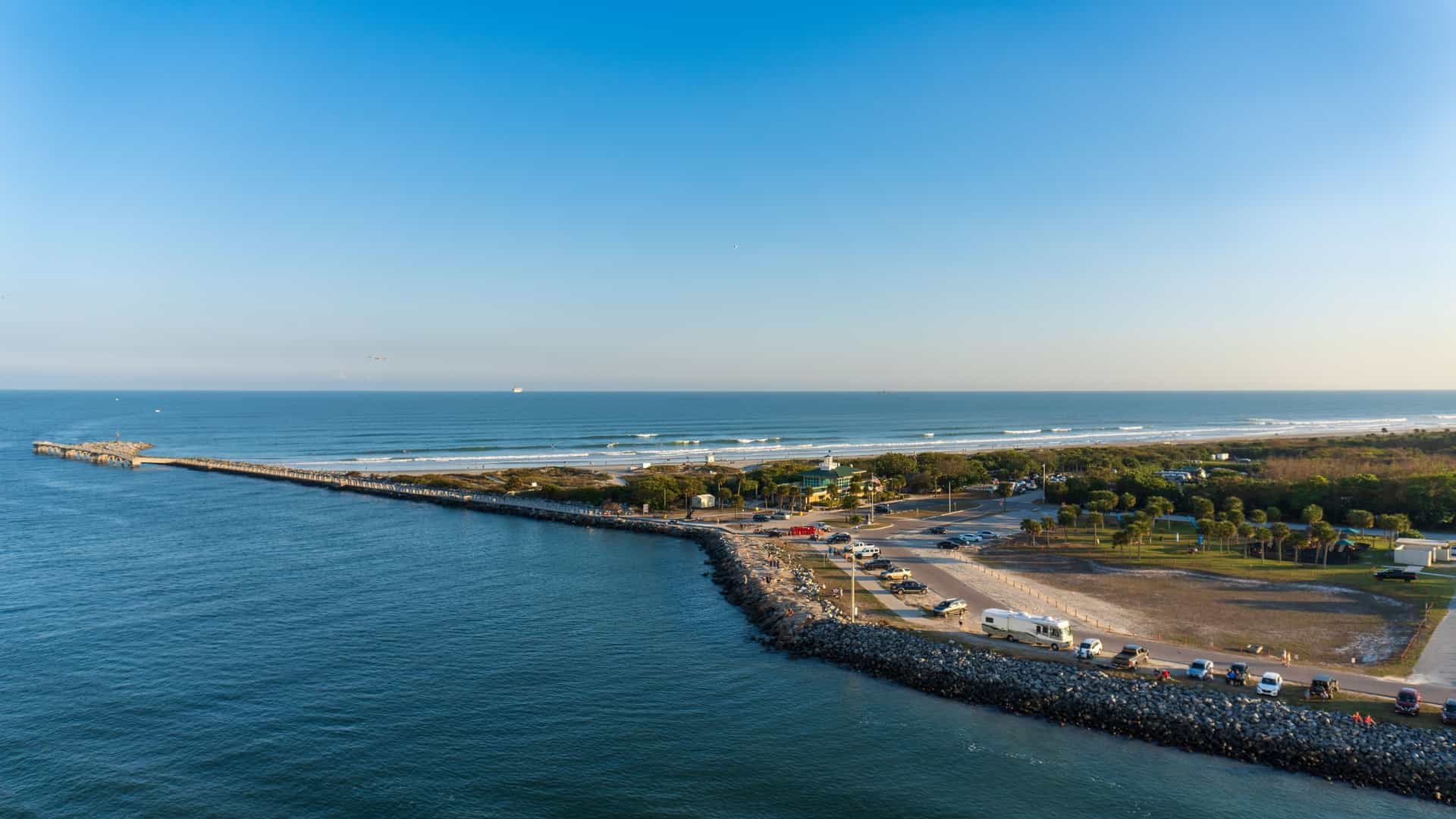 A beautiful day at Jetty Park in Port Canaveral, Florida, with the paved fishing pier extending out into the blue water of the Atlantic Ocean. Palm trees and green grass line the park's beach, and a cruise ship is visible in the distance.