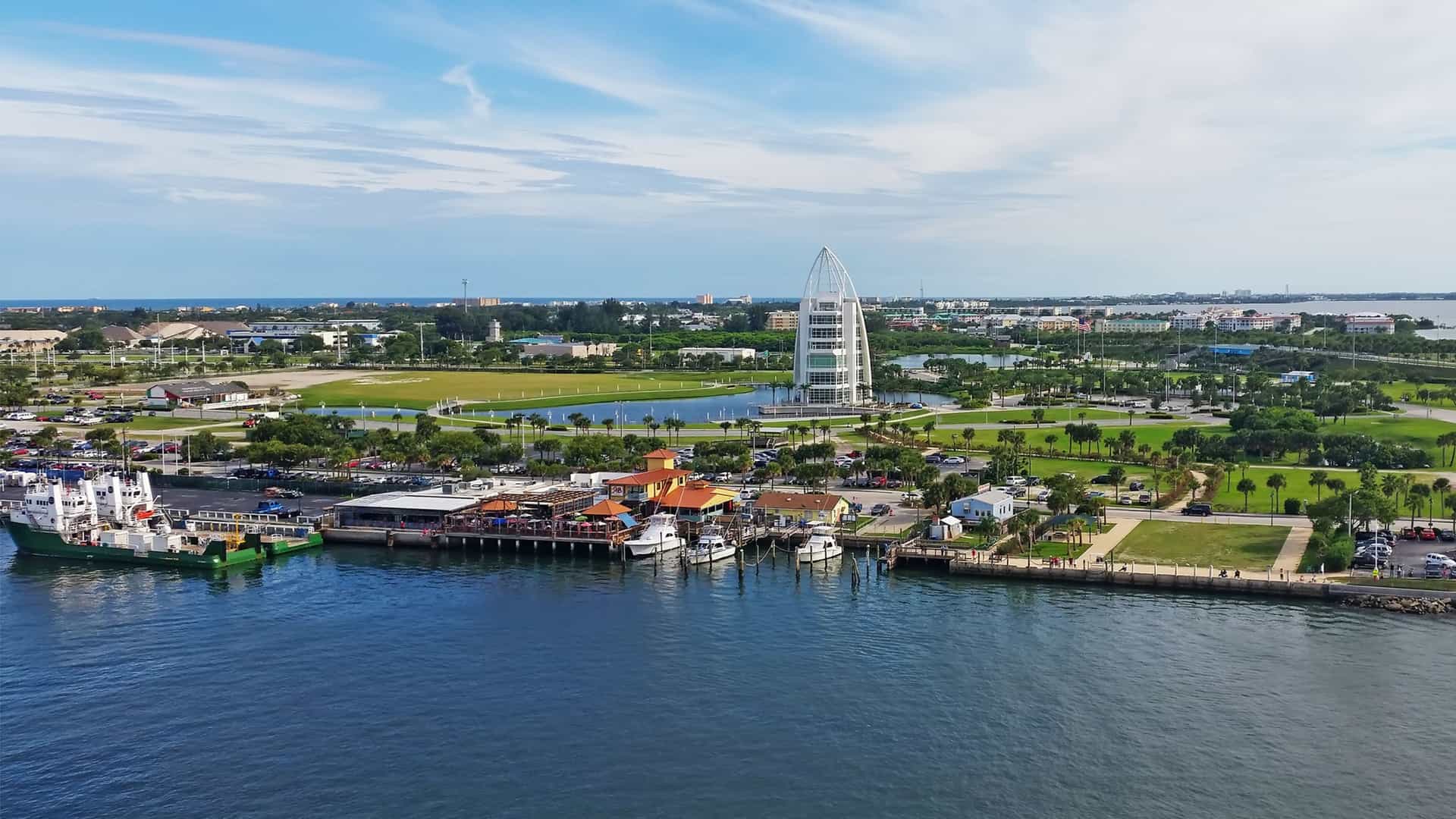 The seven-story Exploration Tower, a modern architectural landmark at Port Canaveral, Florida. Its distinctive, sloping, sail-like design features an iridescent, color-changing metal exterior and exposed steel framework, situated against a waterfront backdrop.