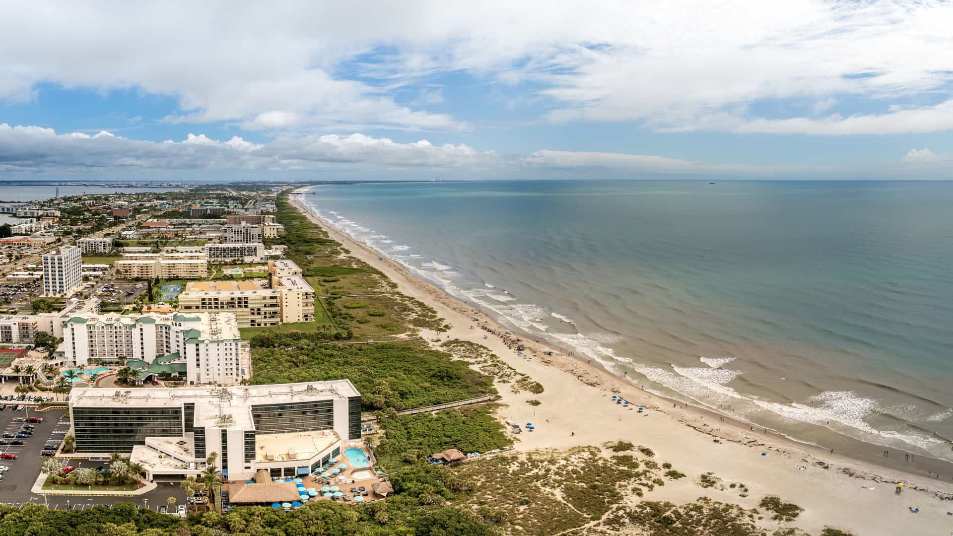 An aerial panoramic view of Cocoa Beach, Florida, showing a long stretch of sandy coastline and the Atlantic Ocean. The image features high-rise hotels and condominiums lining the beach, dense urban development on the barrier island, and the dark blue water of the Banana River on the western side.