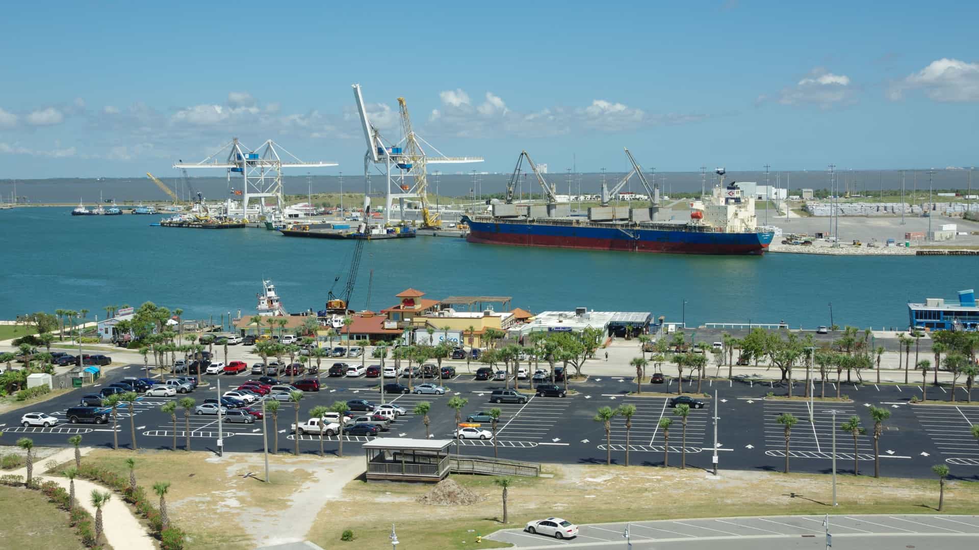An aerial view of the industrial harbor at Port Canaveral, Florida, showing a large cargo ship docked alongside the pier. The foreground features a vast, empty asphalt parking lot, while the background is dominated by large, white gantry cranes used for handling cargo.