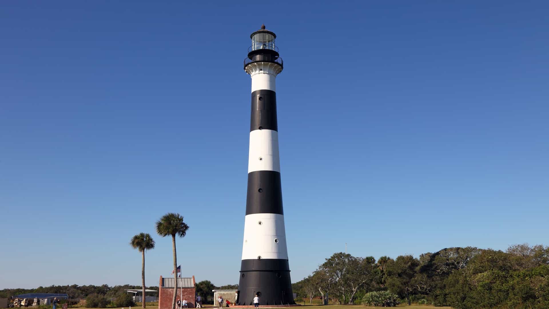 The Cape Canaveral Lighthouse, a tall, conical cast-iron tower with alternating black and white stripes, stands against a clear sky in Florida, USA. The historic 1868 lighthouse, which is 151 feet tall, is situated on the restricted grounds of the Cape Canaveral Space Force Station.