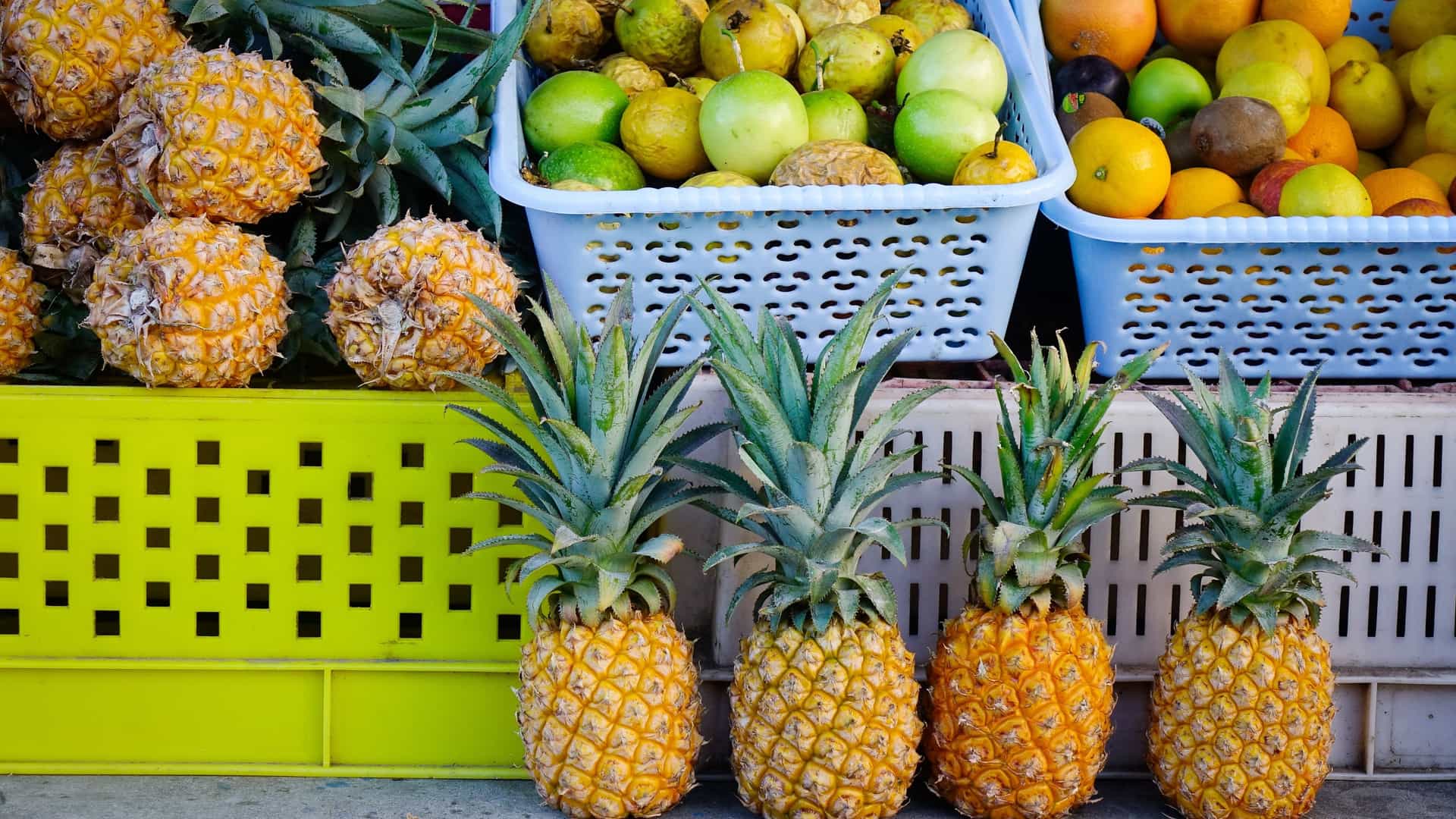 A vibrant fruit stall displaying rows of ripe, whole pineapples in the foreground, with baskets of passion fruit, guavas, and oranges stacked behind them. This scene captures the colorful fresh produce section of the Central Market in Port Louis, Mauritius.