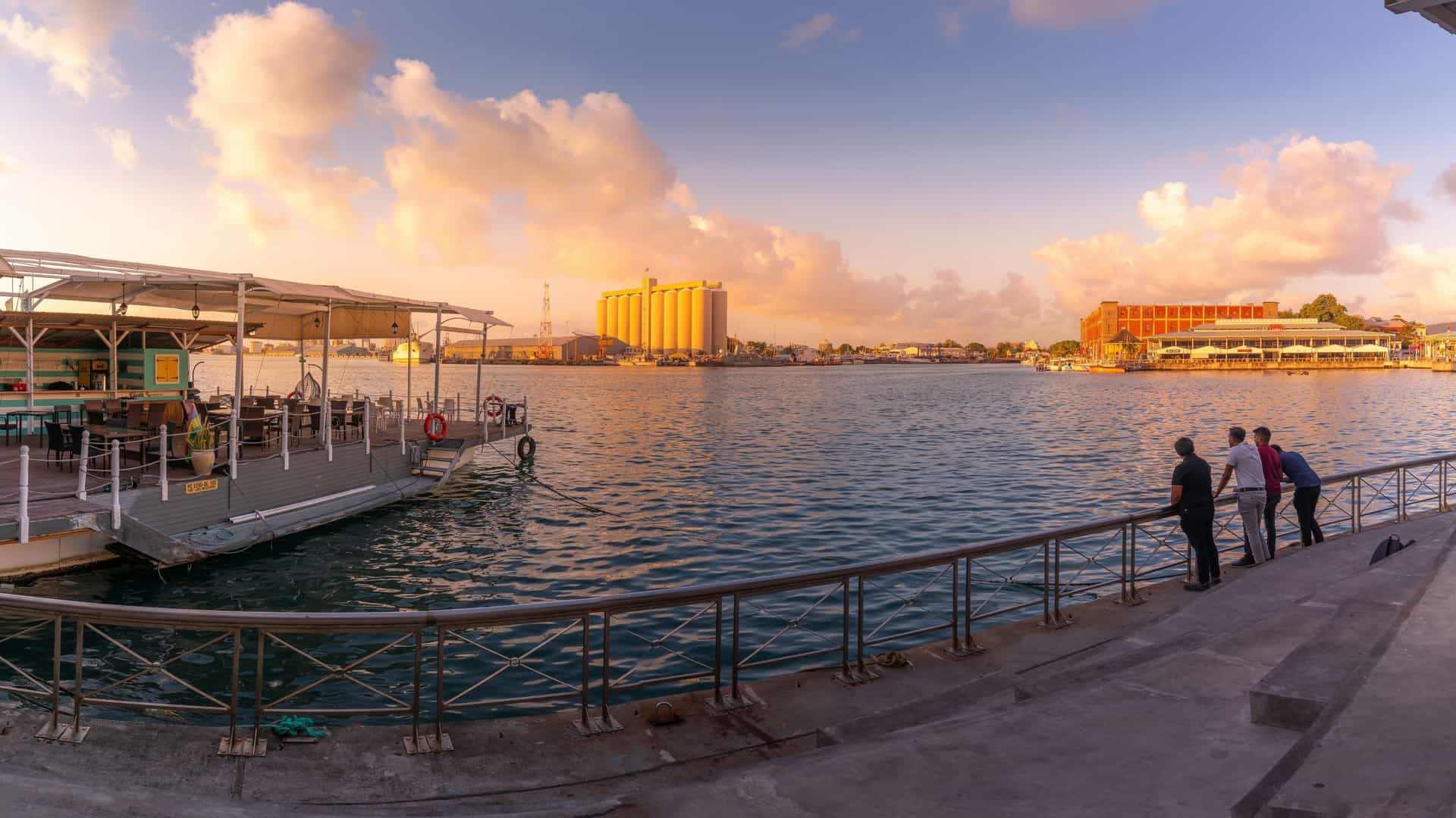 A scenic view of the Caudan Waterfront in Port Louis, Mauritius, at sunset. The image shows the modern buildings and the harbor along the waterfront, with lights beginning to twinkle. The sky is filled with the warm colors of the sunset, and a mountain range is visible in the background, completing the urban and natural landscape.