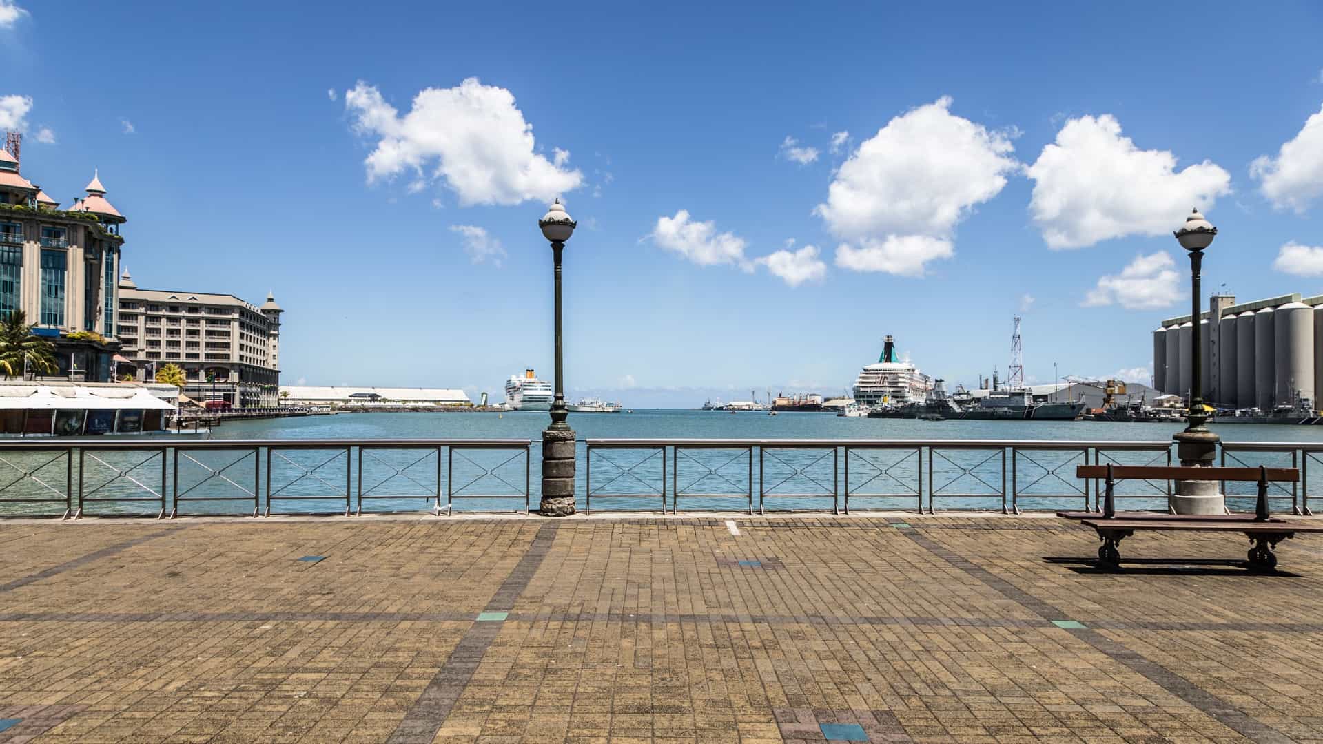 A bright, panoramic photograph of the modern Port Louis waterfront promenade in Mauritius. The foreground features a bustling boardwalk lined with palm trees and modern buildings, including the distinctive, umbrella-covered area of the Caudan Waterfront complex. The harbor is visible to the right, with various ships and the distant mountains surrounding the capital city.