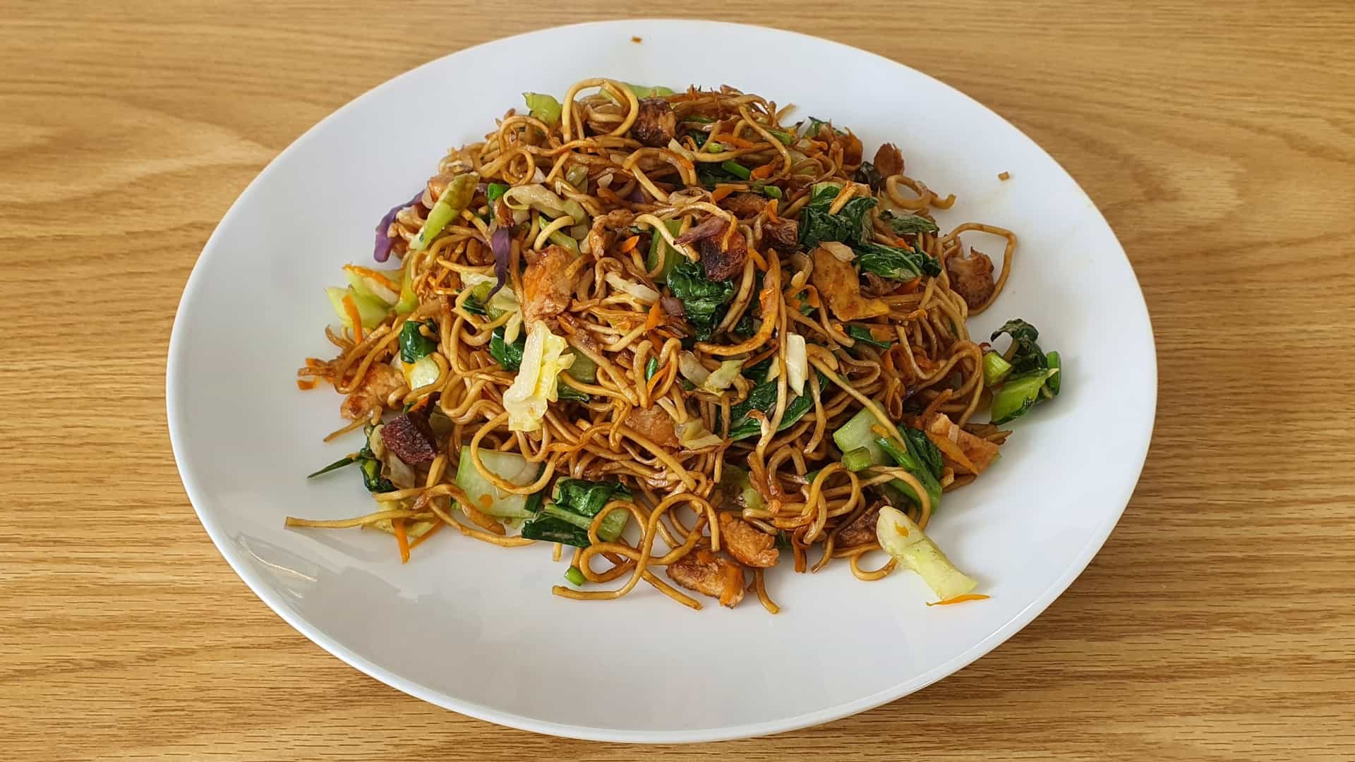 A close-up of a plate of Mine Frire (Mauritian fried noodles), a local specialty. The colorful dish features thick yellow noodles stir-fried with small shrimp (crevettes), chunks of chicken, chopped green vegetables (such as bok choy or scallions), and topped with a fried egg. The dish is presented on a simple white plate.