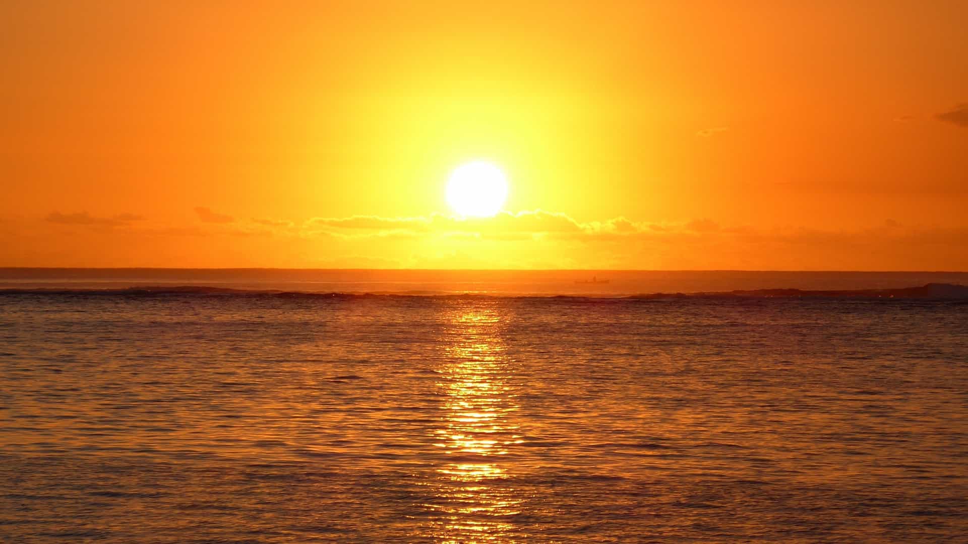 A breathtaking photograph of the sunset over the lagoon near the beaches of Le Morne, Mauritius. The sky is ablaze with warm orange, yellow, and purple hues reflecting on the calm, shallow water. In the background, the distinctive, rugged silhouette of Le Morne Brabant mountain rises prominently, dominating the horizon.