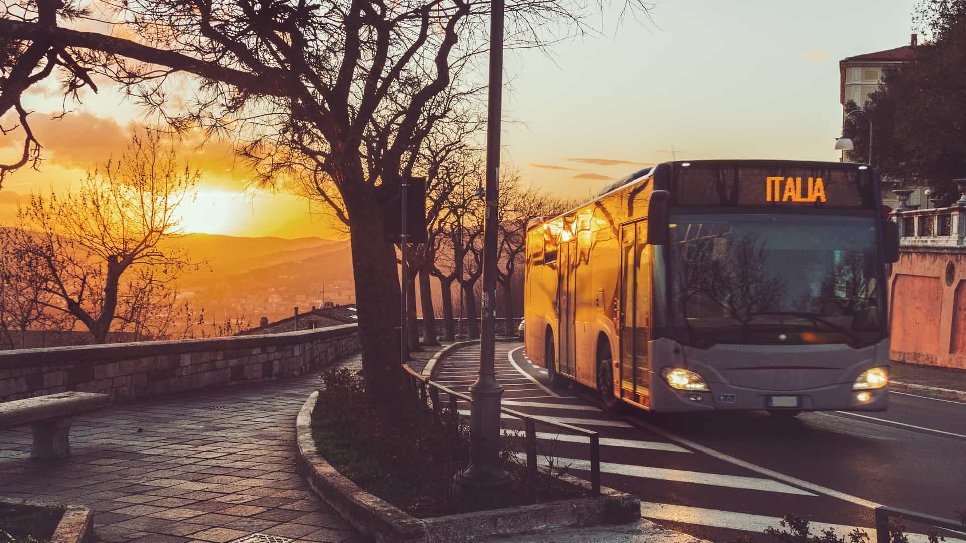 A dramatic photograph taken at sunset on a hillside road in Italy. A large passenger bus, which could function as a shuttle bus to Piazzale Roma, is illuminated by the warm, golden light of the setting sun as it drives around a curve. Bare tree branches frame the scene, and a stone wall lines the sidewalk. The destination sign on the bus clearly reads "ITALIA," with a scenic view of rolling hills in the background.