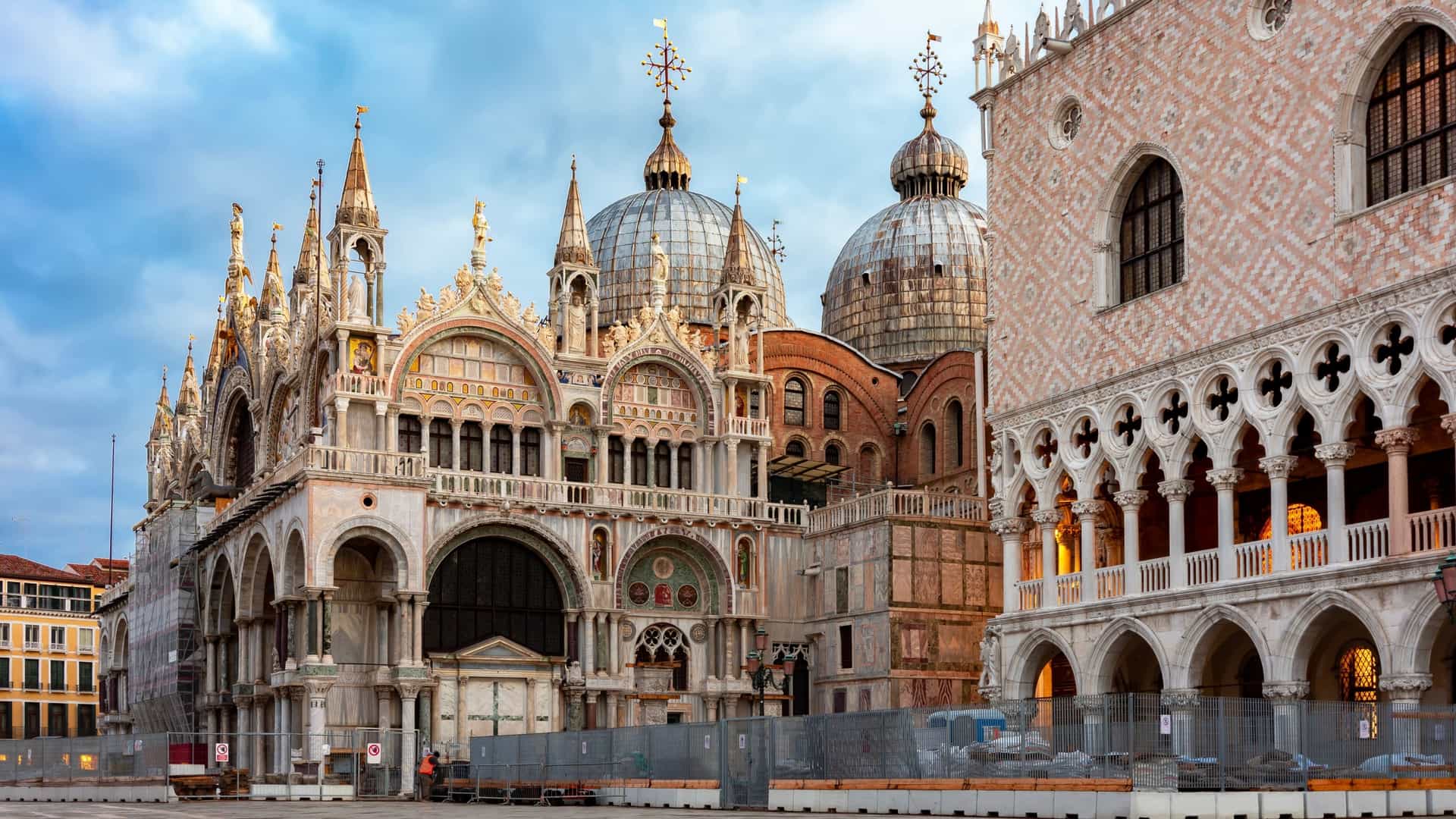 A photograph capturing the ornate, Byzantine-influenced facade of St. Mark's Basilica (Basilica di San Marco) in Venice, Italy. The upper section displays four large, ornate bronze horses and the arched mosaics above the porticos. The scene is bright and highlights the intricate marble work and domes of the historic cathedral.