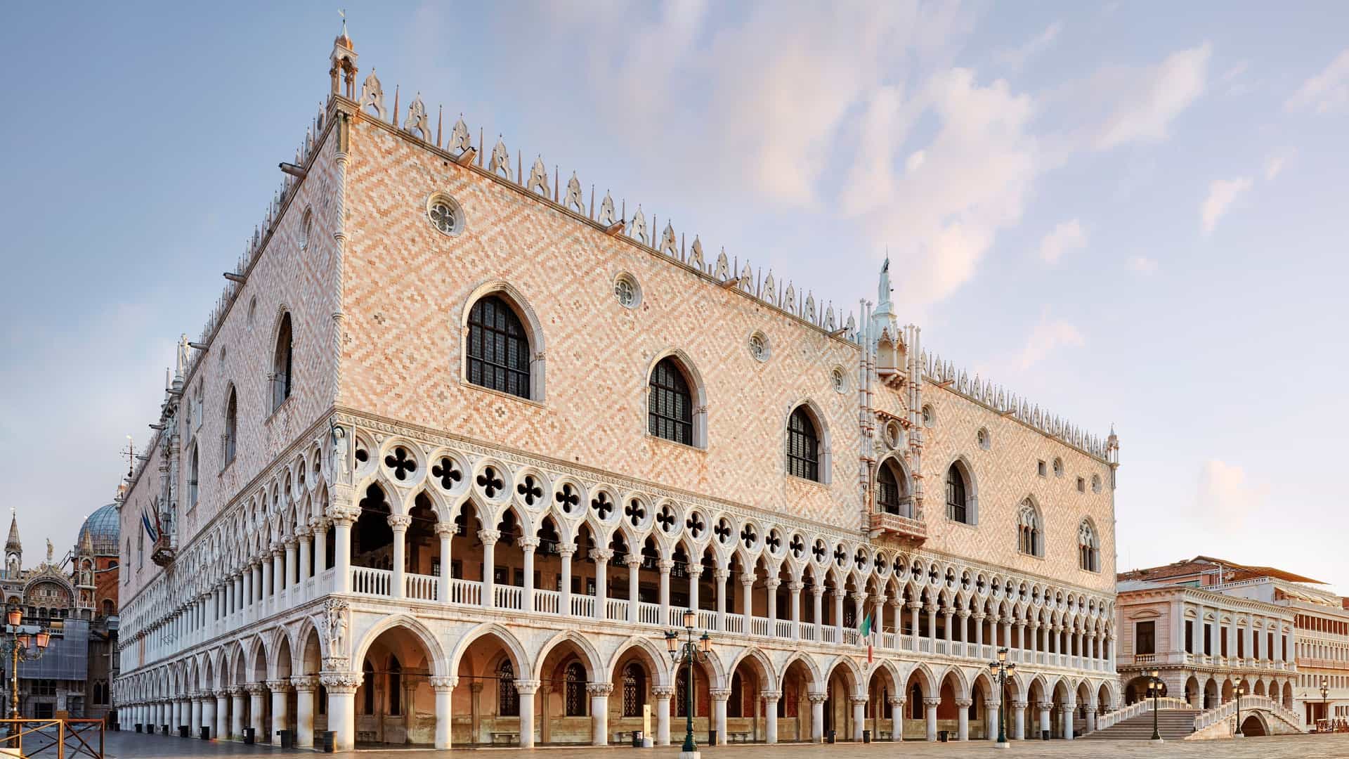 A bright photograph of the magnificent, Gothic Palazzo Ducale (Doge's Palace) in Venice, Italy. The palace is characterized by its white and pink diamond-patterned marble facade and a series of delicate, arched loggias on the upper and lower levels. The historic structure stands prominently along the waterfront, with a section of the Venetian lagoon visible in the foreground.
