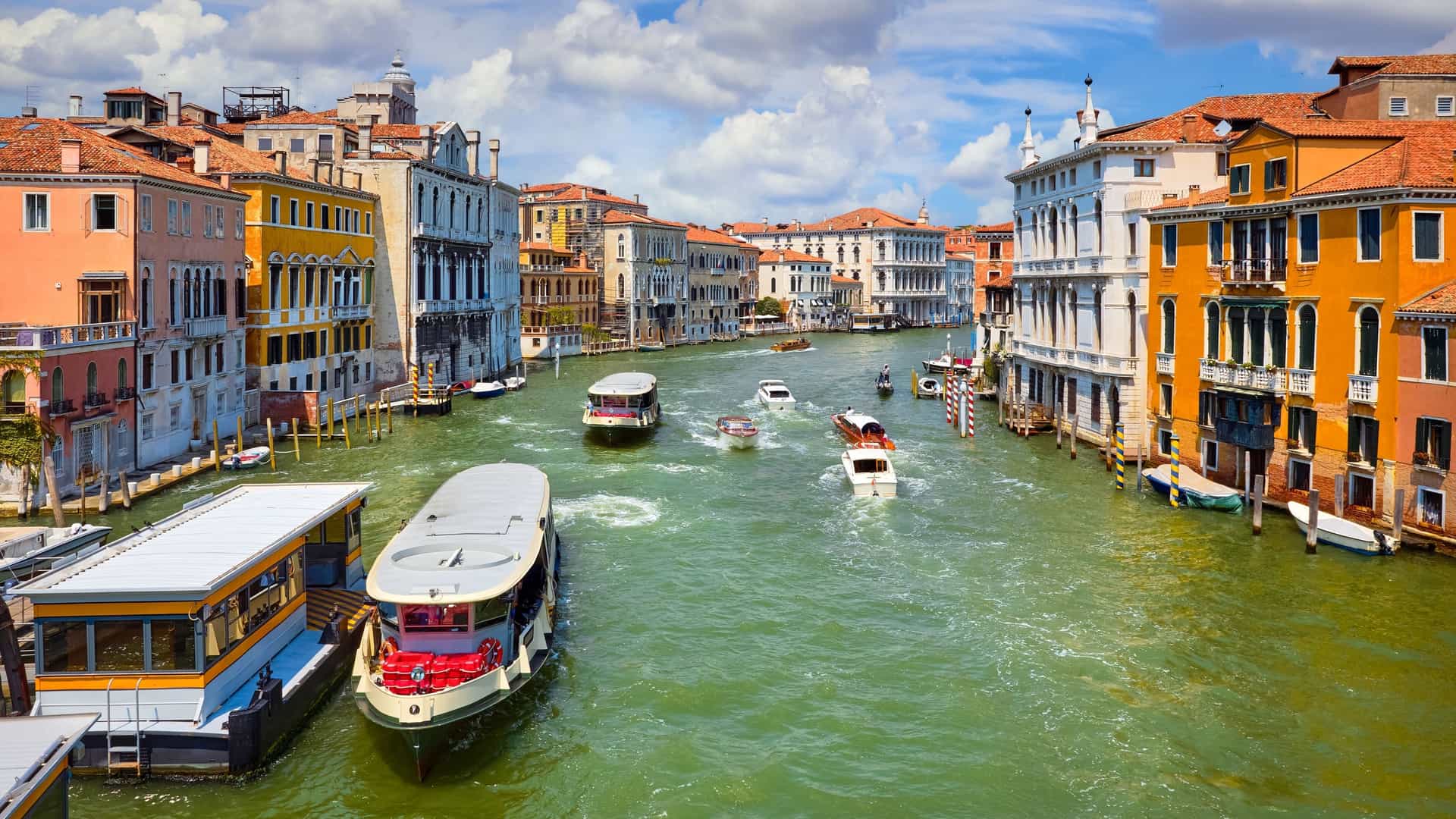 A bright, sunlit photograph of the Grand Canal in Venice, Italy. The canal is wide and features greenish-turquoise water with several boats and water taxis moving along it, offering the perfect opportunity to Ride a Vaporetto on the Grand Canal. The banks are lined with colorful, historic Venetian palaces in shades of orange, yellow, and white, with the dense urban landscape filling the background under a blue sky with scattered clouds.