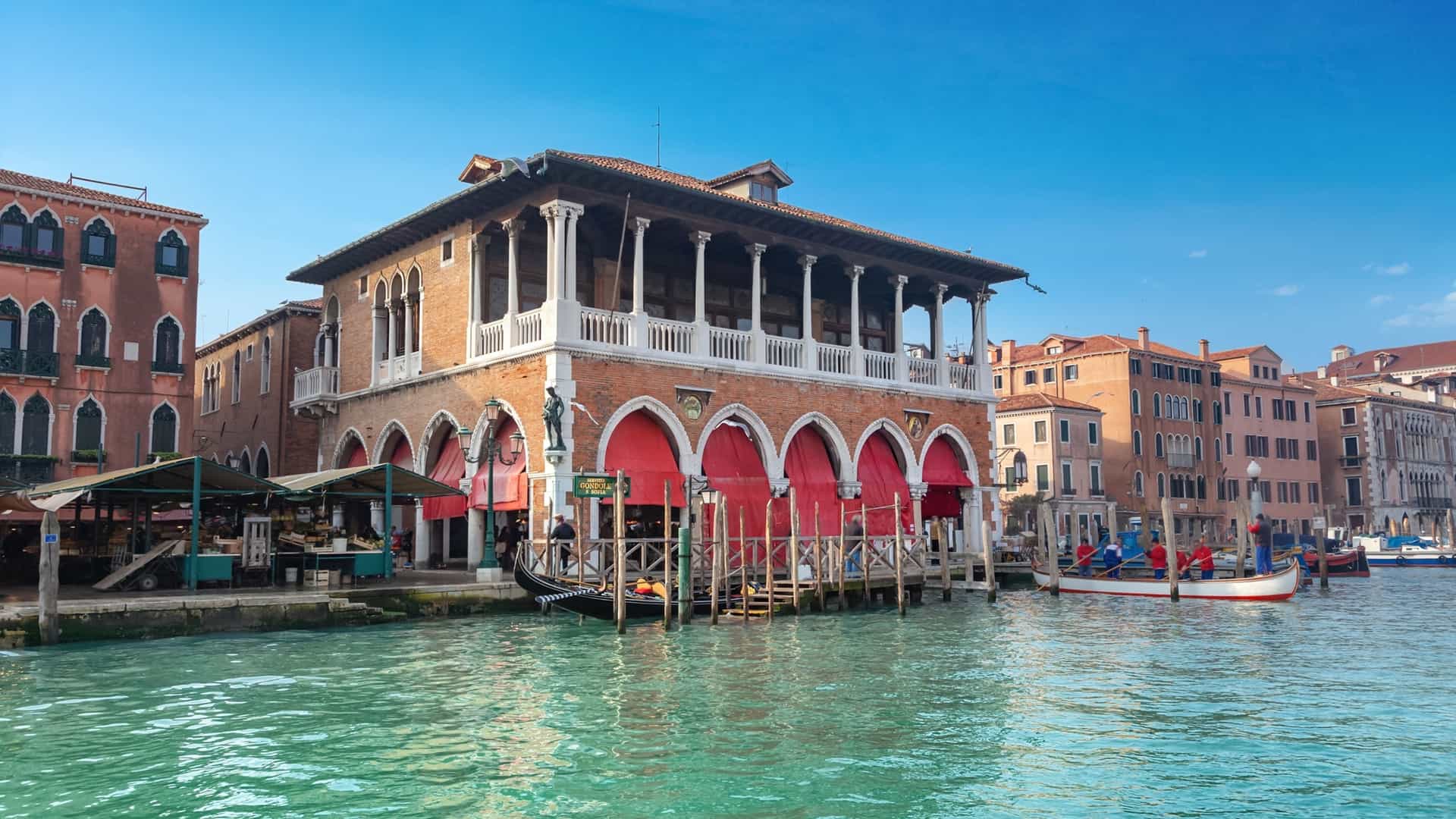 A bright, bustling photograph of the Rialto Fish Market (Pescheria) in Venice, Italy. The market is situated under a historic, neo-Gothic stone arcade. Several vendors are visible behind wet, tiled stalls displaying a variety of fresh seafood and fish laid out on ice. The scene captures the lively atmosphere of one of Venice's oldest markets.