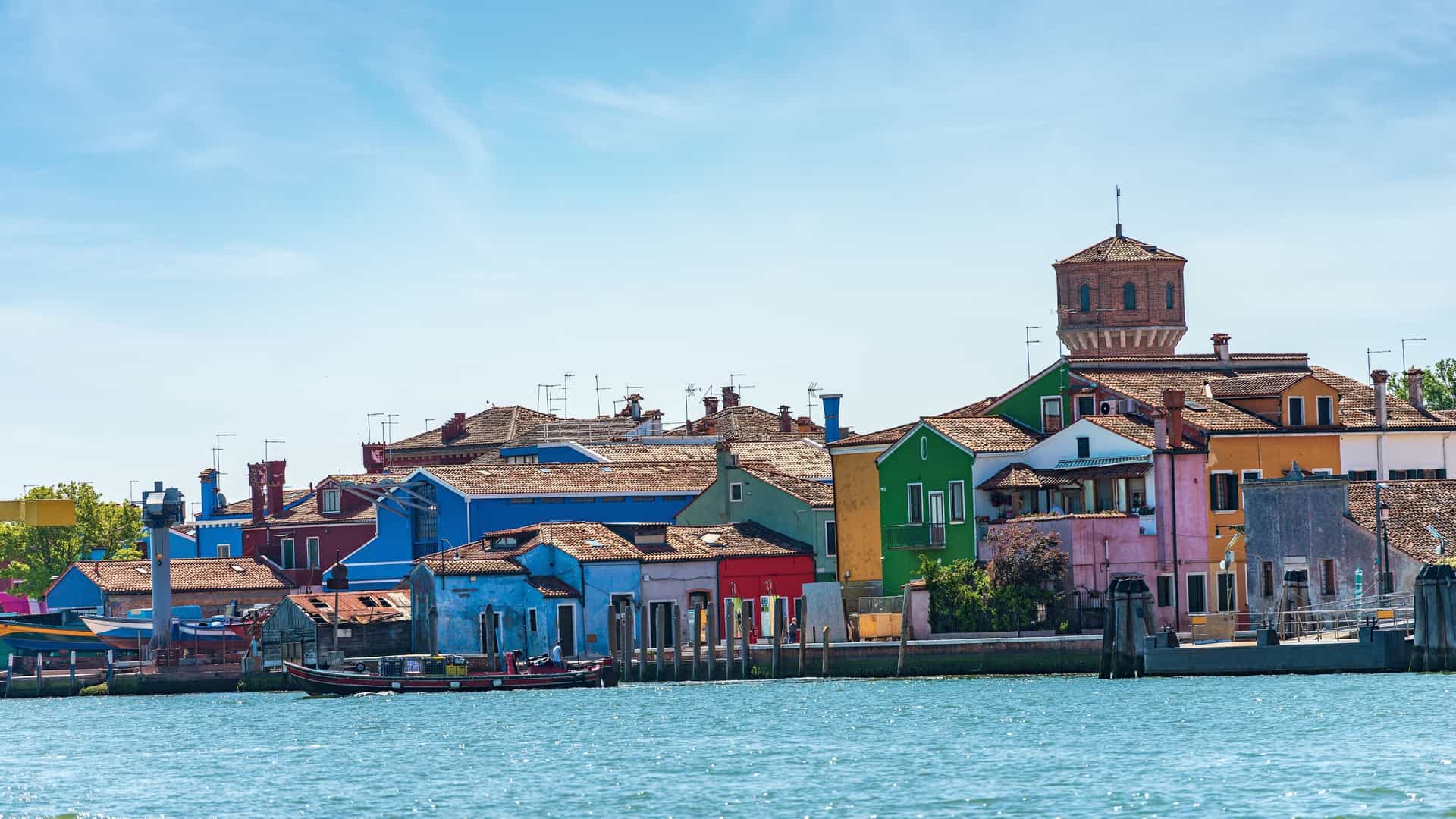 A highly vibrant photograph of the characteristic, multi-colored houses lining a small canal on Burano Island, near Venice, Italy. The houses are painted in brilliant shades of pink, blue, yellow, and red. The calm water of the canal reflects the colorful buildings, and a small wooden bridge crosses the waterway in the foreground under a bright blue sky.