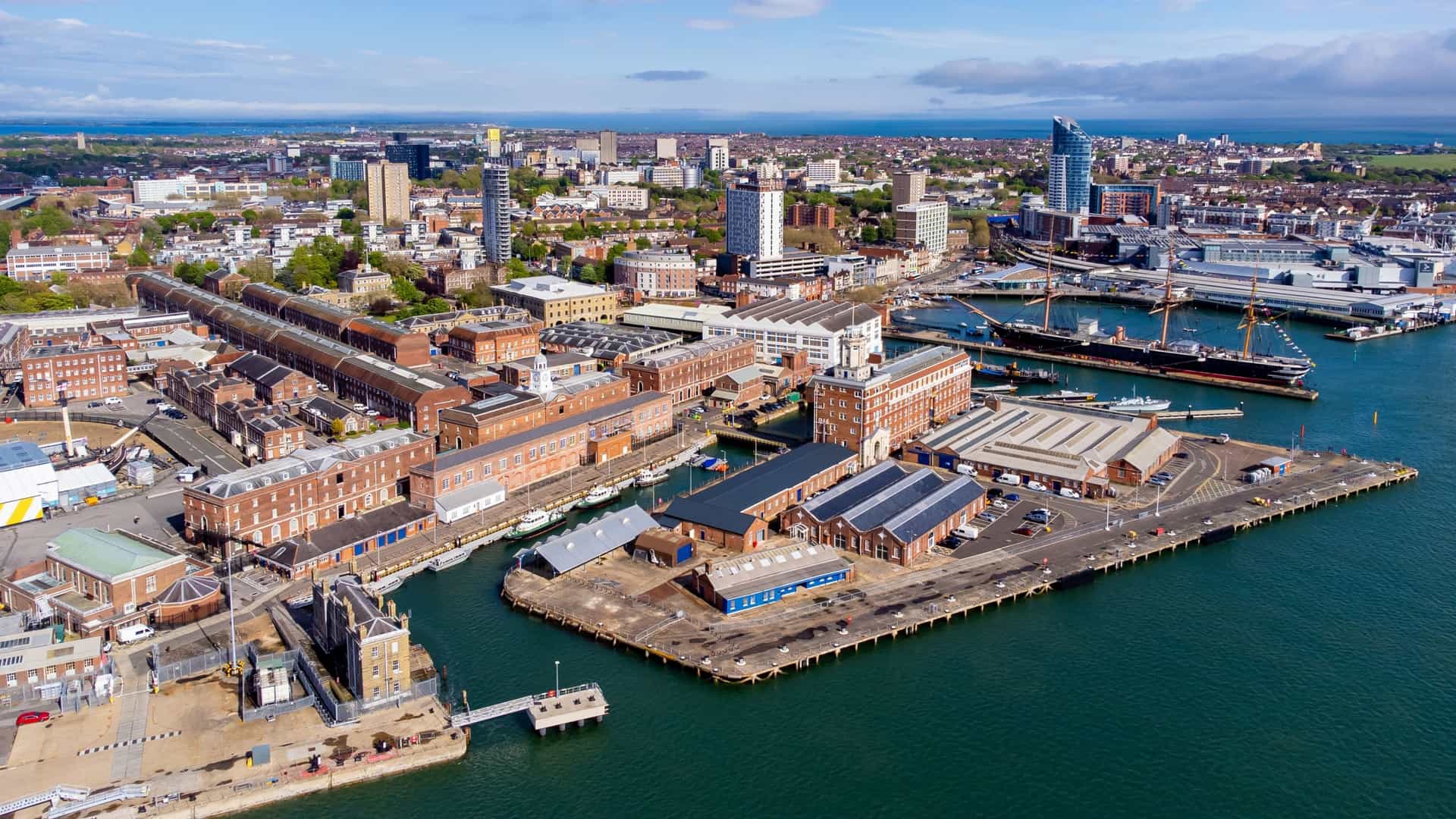 An aerial view of Portsmouth Historic Dockyard and the active Royal Navy Base in Portsmouth, England. The image captures several famous historic warships docked in the harbor, including the ironclad frigate HMS Warrior (1860) and potentially the wooden warship HMS Victory (1759), alongside modern Royal Navy vessels and the nearby landmarks like the Spinnaker Tower in the background.