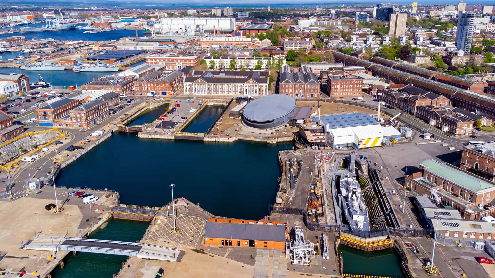 An aerial view of the Mary Rose Museum complex at the Portsmouth Historic Dockyard. The museum, which houses the Tudor warship, is characterized by its large, black, rounded building with a prominent curved roof, positioned centrally on the dockyard floor next to historic water basins and surrounded by other naval buildings.