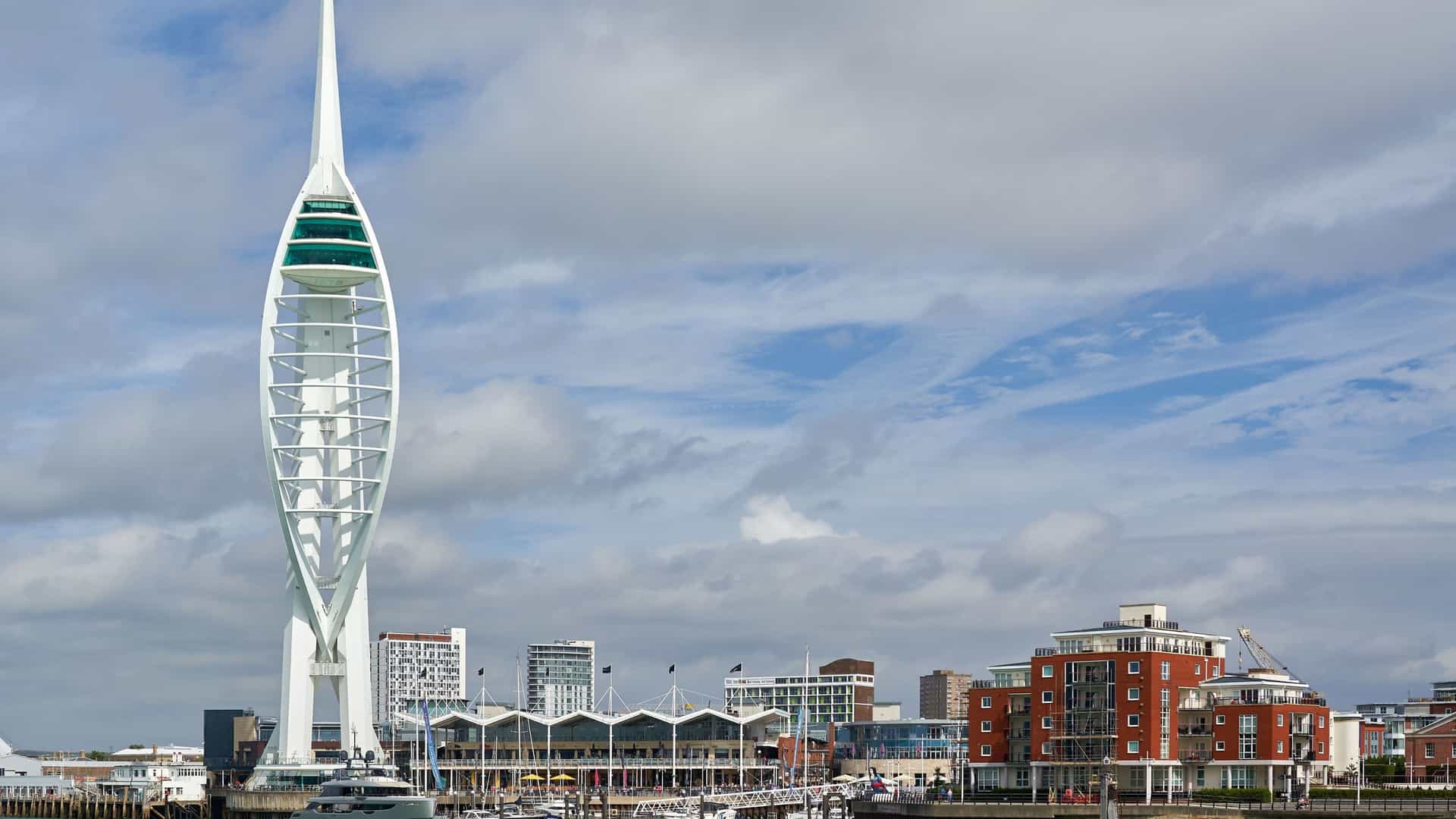 A beautiful sunset view over Portsmouth Harbour and the Gunwharf Quays area in England. The image is dominated by the distinctive white sail-shaped structure of the Spinnaker Tower, illuminated by the warm light of the setting sun, with the city's waterfront, including the Portsmouth Historic Dockyard, visible below.