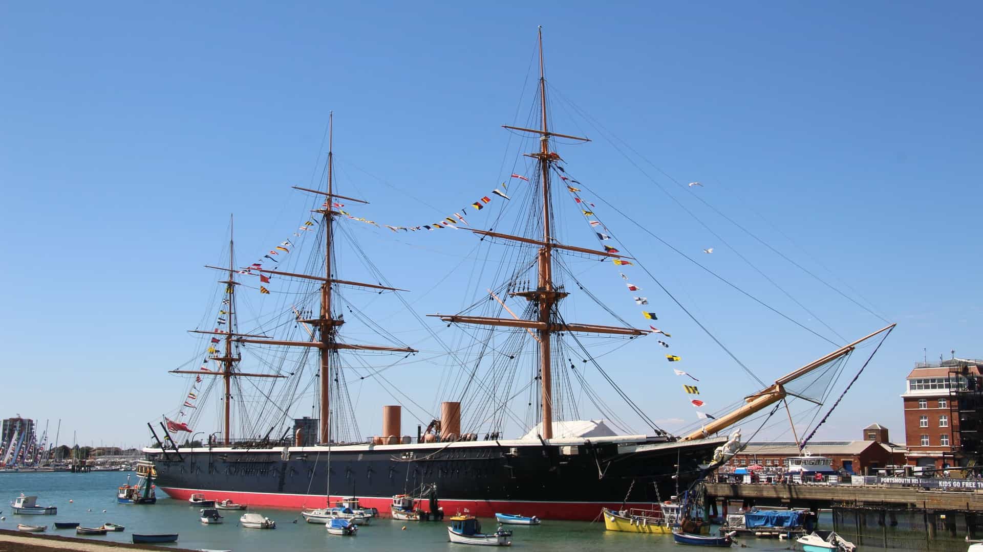 A full-length view of the HMS Warrior (1860), Britain's first iron-hulled, armored battleship, moored in the water at Portsmouth, England. The black-hulled Victorian museum ship is shown from a low angle, emphasizing its massive size, masts, and rigging, with the buildings of the historic dockyard visible in the background.