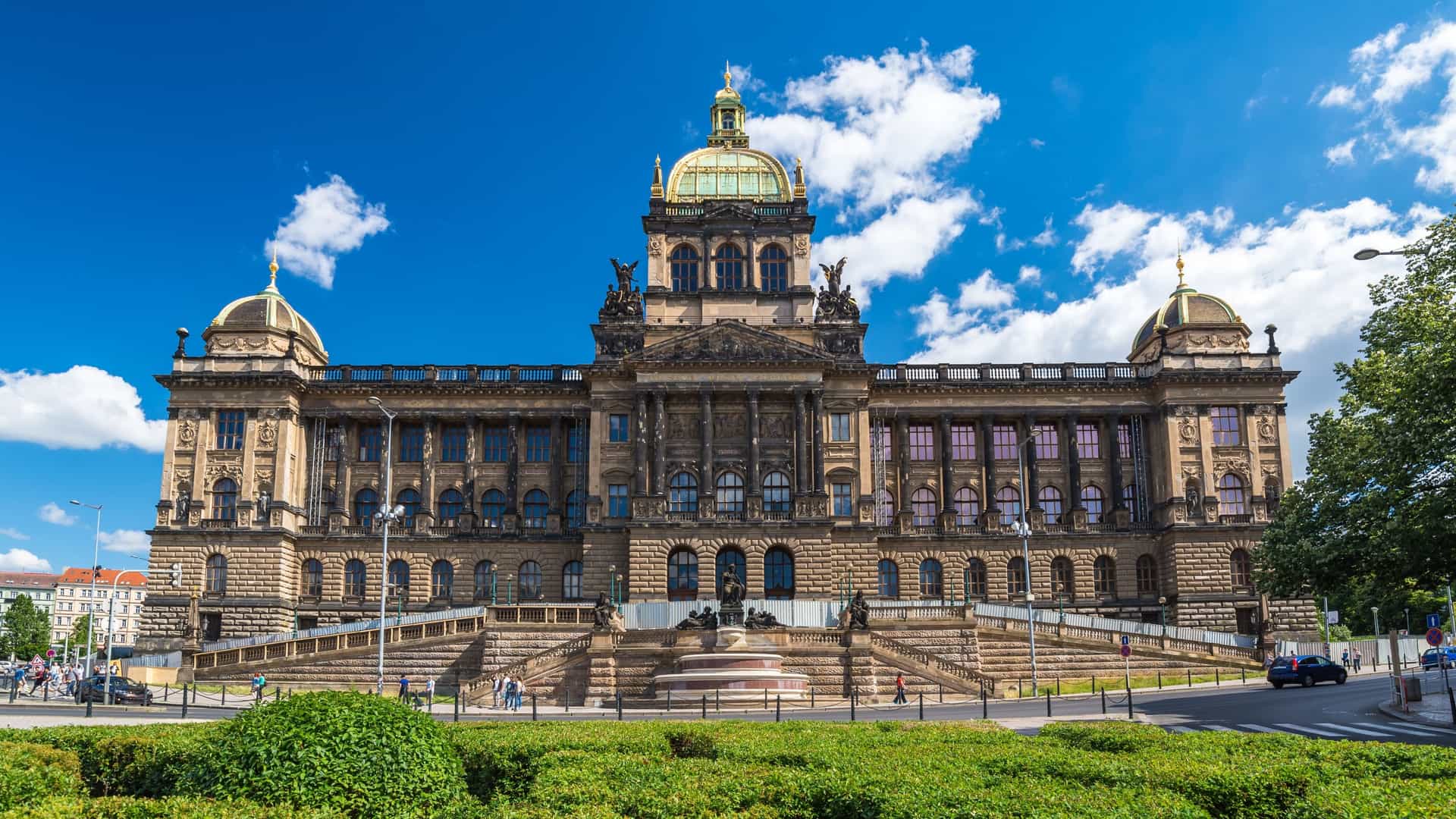 A wide, slightly elevated view of Wenceslas Square (Václavské náměstí) in Prague, Czech Republic, showing the long boulevard lined with historic buildings, with the grand, Neo Renaissance National Museum building visible at the far, uphill end. The square is bustling with pedestrians and traffic.