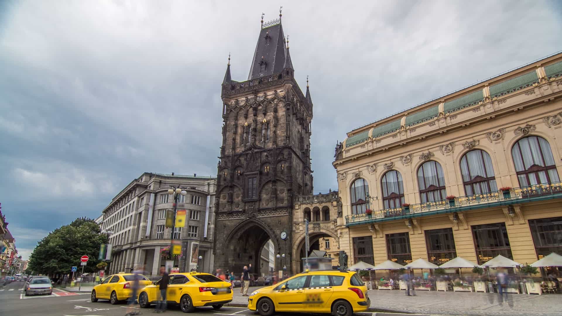 A view of the Gothic Powder Tower (Prašná brána) in Prague, Czech Republic, positioned directly next to the ornate Art Nouveau Municipal House (Obecní dům). The image, which is a timelapse/hyperlapse frame, highlights the contrast between the medieval, dark stone tower and the brightly decorated, domed Art Nouveau building. The scene is set on Republic Square (Náměstí Republiky) and often includes movement blur (hyperlapse/timelapse effect).