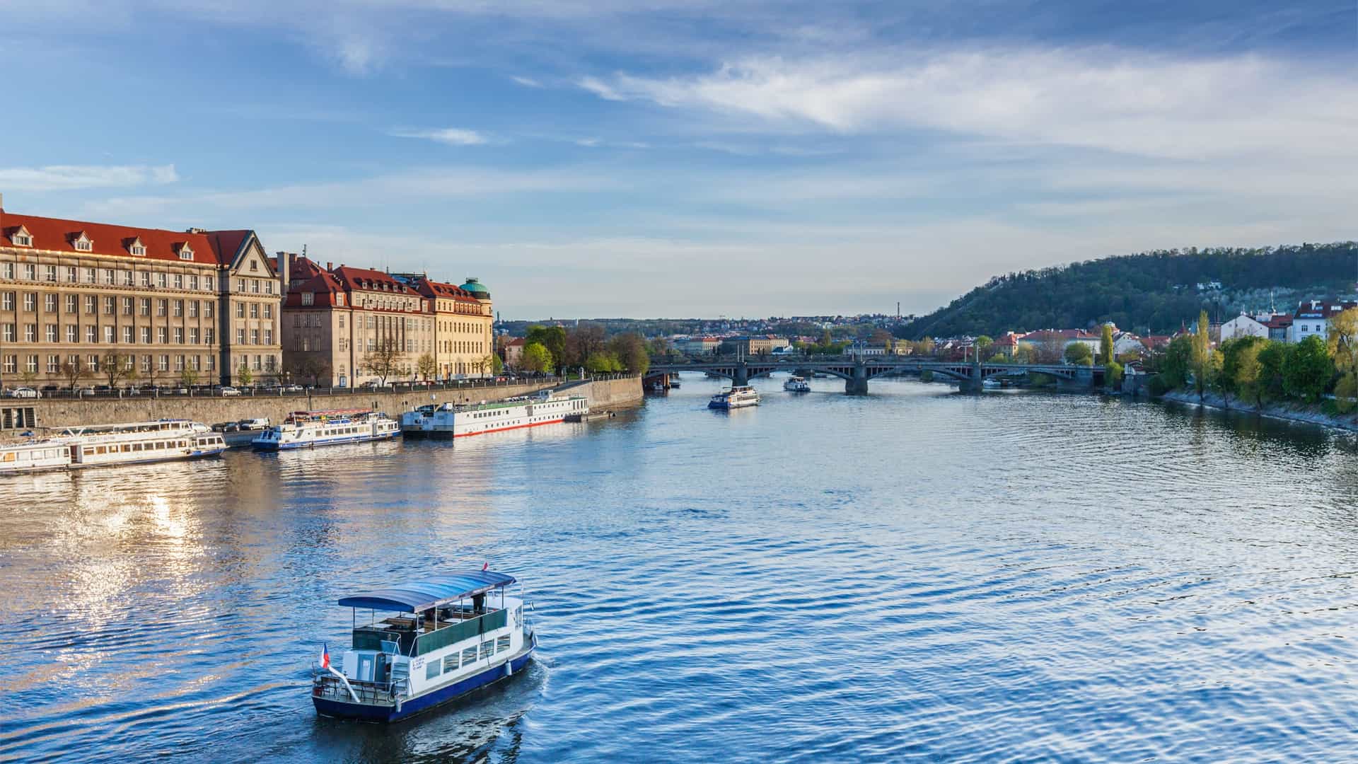 A view of sightseeing or tourist boats navigating the Vltava River in Prague, Czech Republic. The image typically captures the historic cityscape from the water, often featuring famous landmarks like the Charles Bridge, Prague Castle towering on the hill, and the historic buildings along the riverbanks, showcasing the popular river cruise experience.