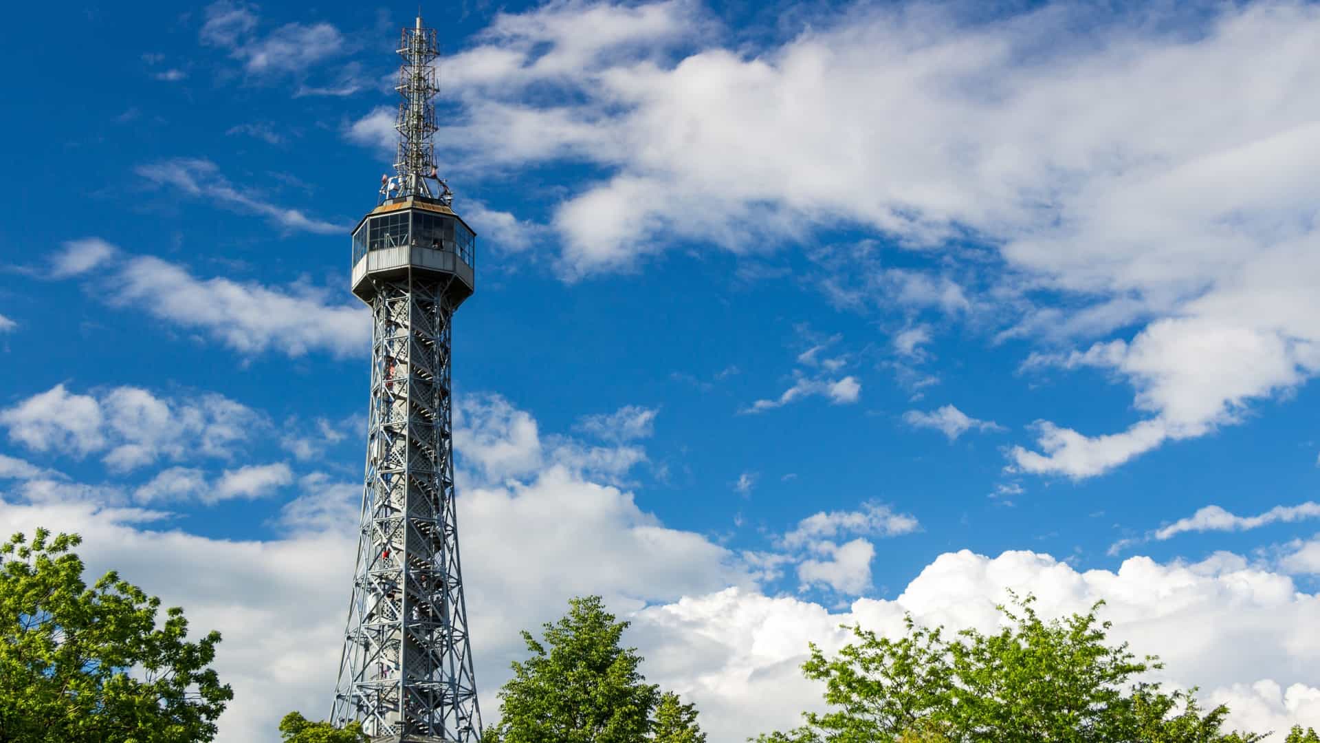 A photograph of the Petřín Lookout Tower in Prague, Czech Republic. The tower is a steel frame structure built in 1891, which resembles a smaller version of the Eiffel Tower in Paris. The image typically captures the tower from a distance or an angle that highlights its lattice steel construction and elevated position on Petřín Hill, often with a view of the surrounding park or the Prague skyline in the background.