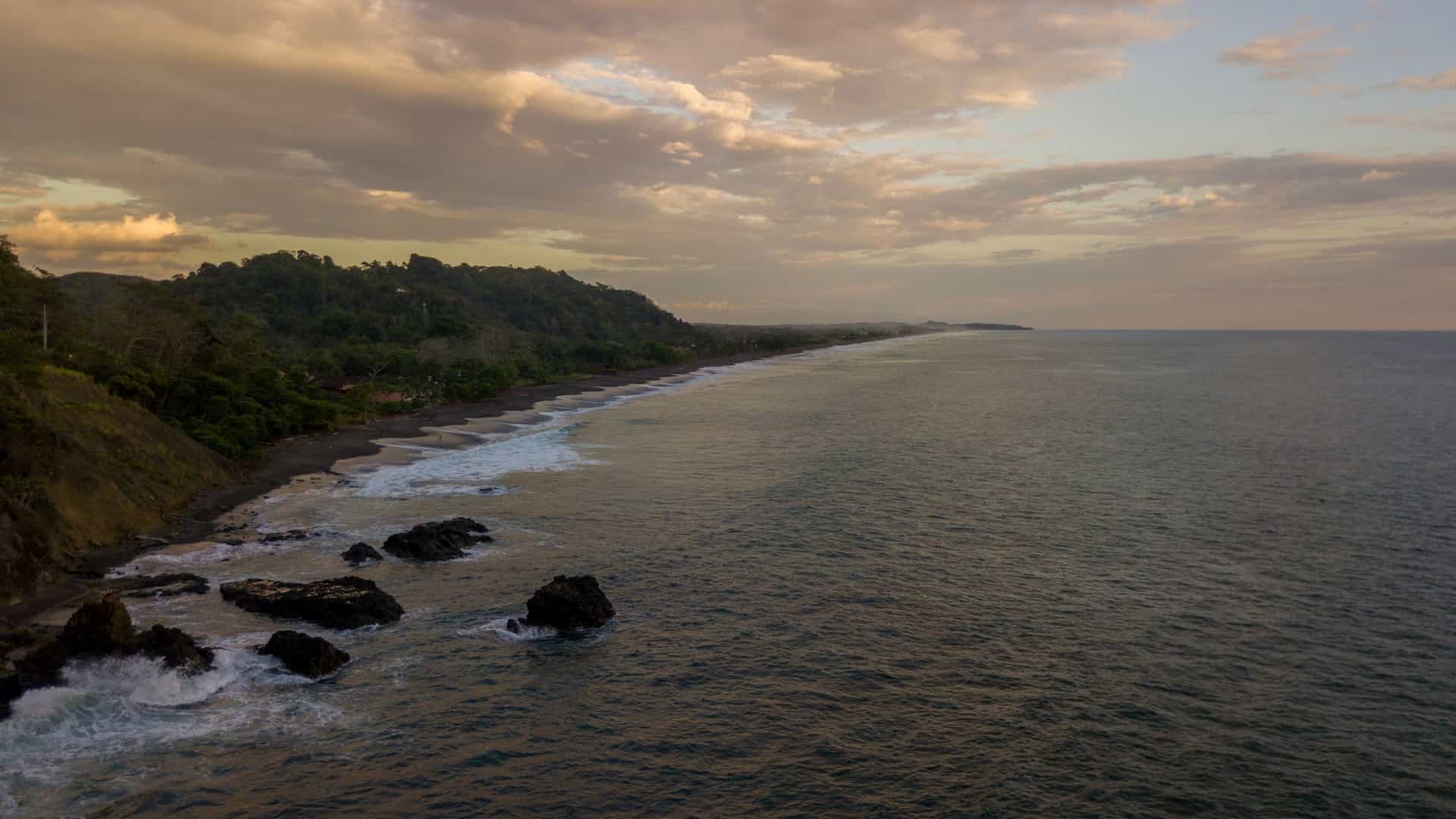 A beautiful aerial view of Playa Hermosa in Guanacaste, Costa Rica. The image captures the curved bay with calm, deep blue water, a stretch of light colored sand beach, and lush green tropical hillsides or mountains framing the coastline. The tranquil bay may feature anchored boats or yachts.