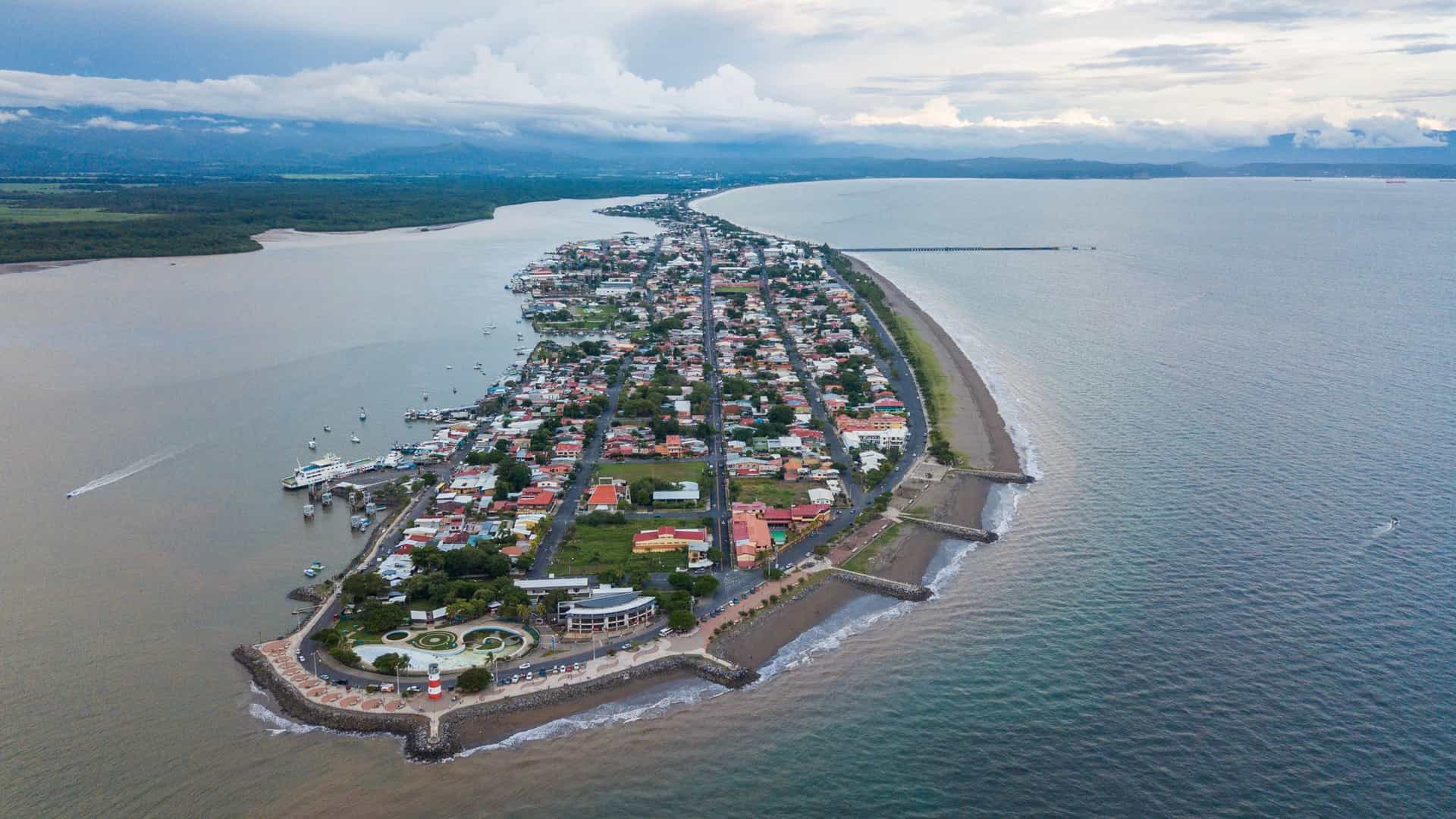A stunning aerial view of Puntarenas, Costa Rica, showcasing the town's unique geography as a narrow peninsula jutting into the Gulf of Nicoya. The densely built town, with low-rise buildings and a long, developed coastline, is clearly visible, flanked by the deep blue ocean on both sides and backed by lush green mountains in the distance. The long, sandy beach and port infrastructure are prominent features.
