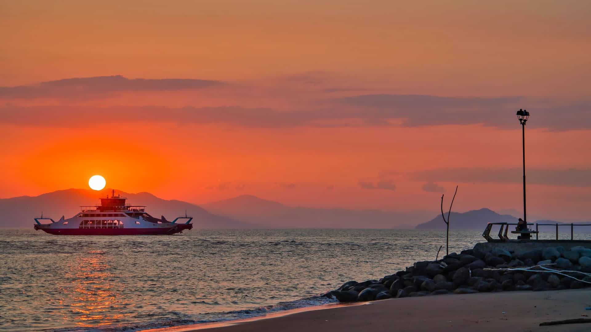 A dramatic photograph of a sunset over the water near Puntarenas, Costa Rica. The sky is filled with intense orange and yellow hues reflecting on the ocean. A large ferry boat is silhouetted against the bright horizon as it moves across the water. The image captures the serene but vibrant end of the day in the Gulf of Nicoya.