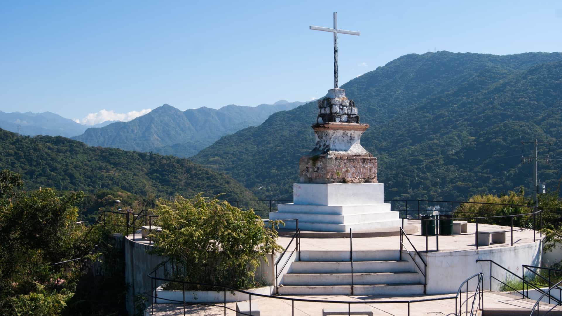 A bright, panoramic photograph of the Mirador Cerro de la Cruz (Hill of the Cross Viewpoint) in Puerto Vallarta, Mexico. In the foreground, a tiered, white stone monument topped with a large, white cross stands on a viewing platform surrounded by railings and steps. The background is dominated by a lush expanse of green, rugged Sierra Madre mountains under a clear blue sky.