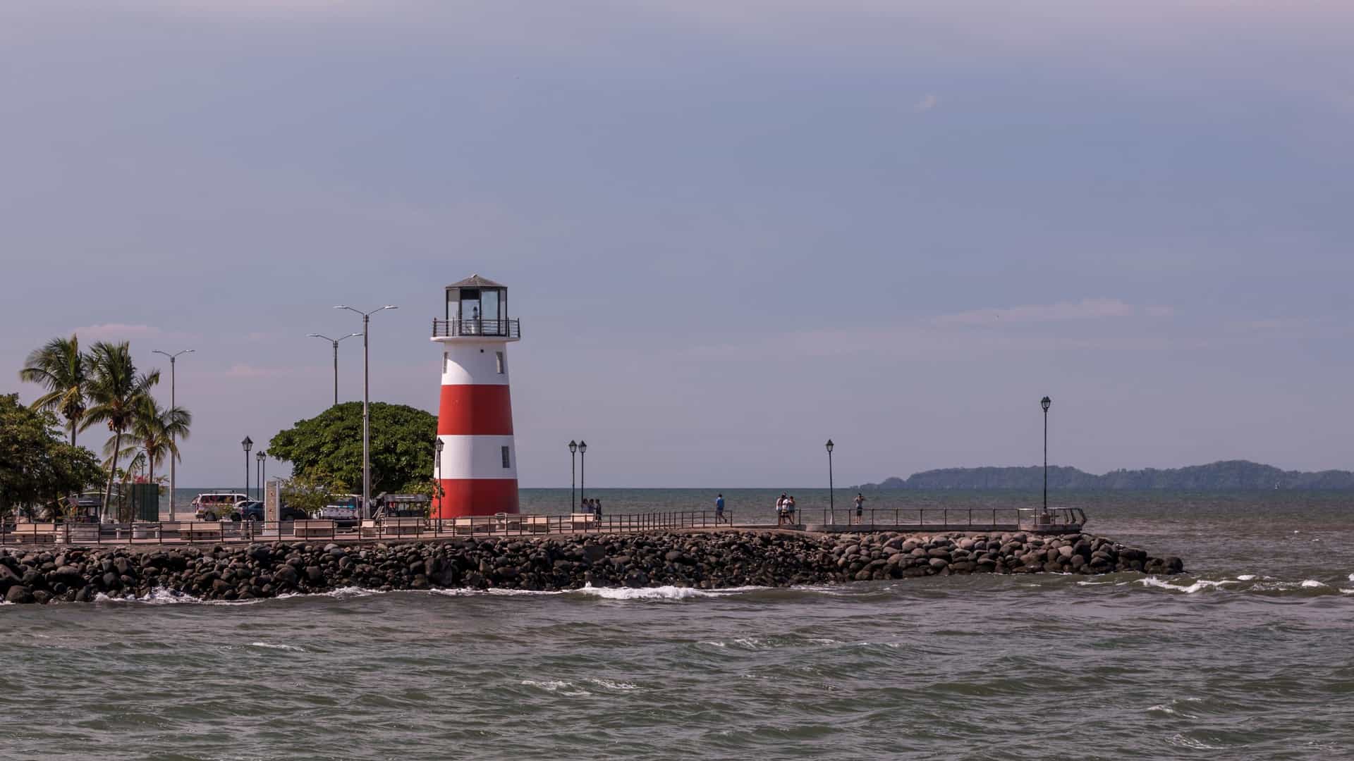 A bright, daytime photograph of the modern, cylindrical, white lighthouse situated on the sandy beach of Puntarenas, Costa Rica. The lighthouse has a distinctive blue spiral railing running up the exterior. It is surrounded by palm trees and overlooks the dark, calm waters of the Pacific Ocean under a blue sky.