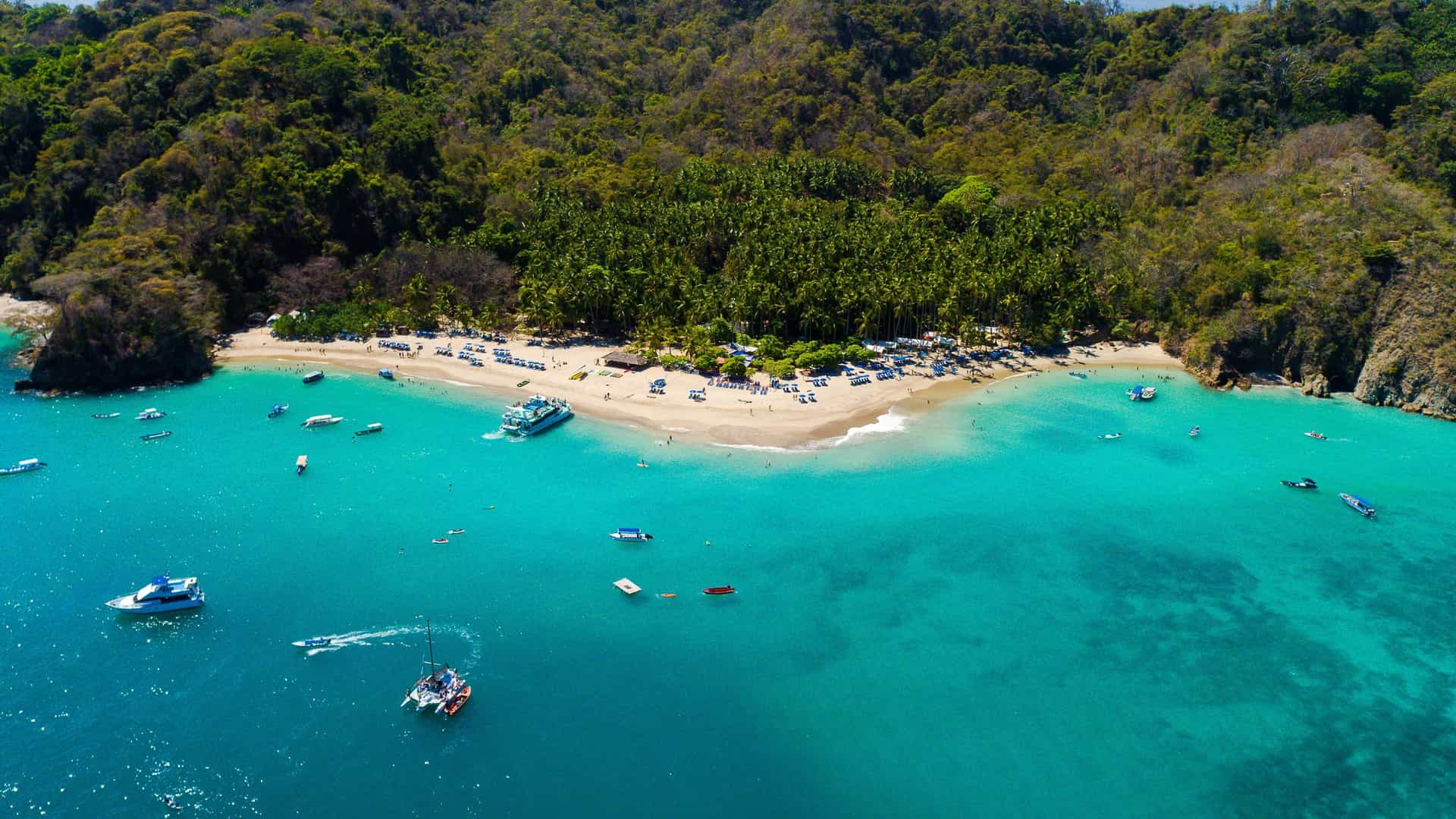 A scenic photograph of the white-sand beach and turquoise water of Tortuga Island, Costa Rica. The beach is crescent-shaped and lined with dense, vibrant green foliage and palm trees. A small group of boats or yachts are anchored offshore in the clear blue water, illustrating a popular tropical tourist destination.