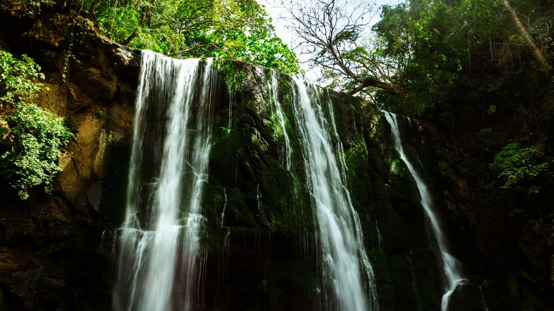 A serene photograph of the El Encanto Waterfall near Esparza, Puntarenas, Costa Rica. A moderate volume of water cascades over a rocky cliff face into a shallow, clear pool at the base. The waterfall is set deep within a lush, green, tropical forest, with surrounding rocks covered in moss and ferns.