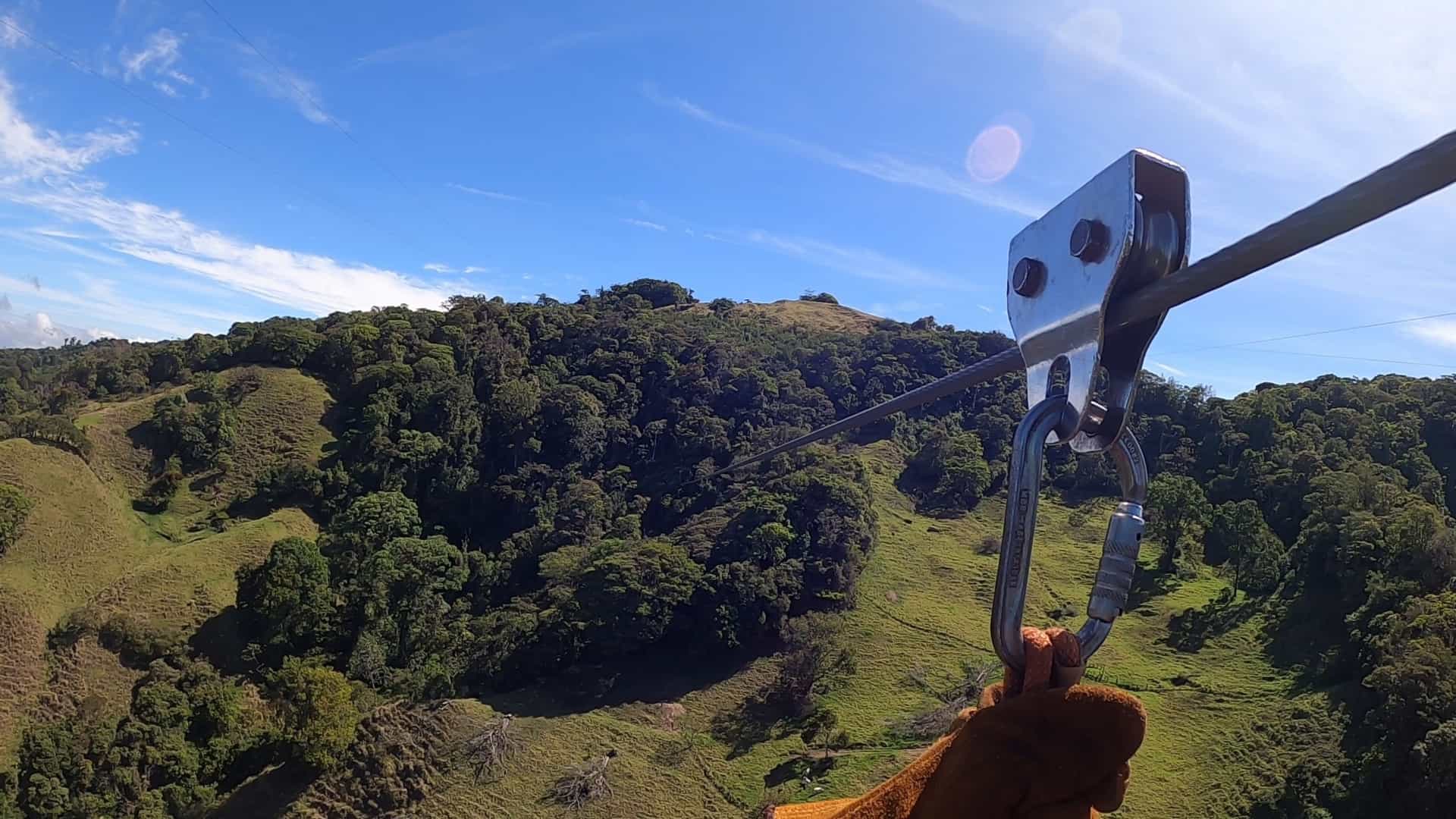A thrilling, high-angle photograph showing a person ziplining across the vast canopy of the Monteverde Cloud Forest in Costa Rica. The person is flying along a cable high above the dense, lush green treetops, with mountains and mist visible in the distance, capturing the adrenaline-pumping nature of the adventure activity.