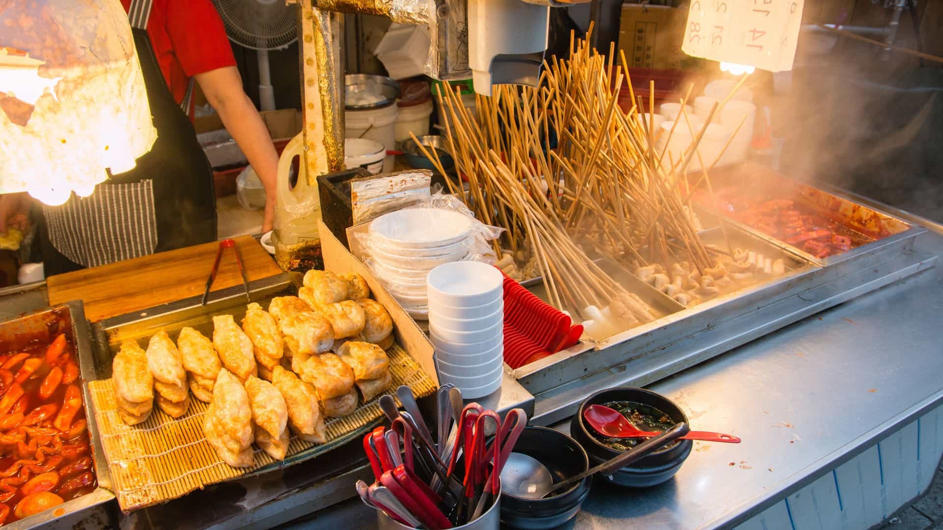 A close-up, vibrant photograph of a popular Korean street food dish, Tteokbokki (cylindrical rice cakes), simmering in a bright red, glossy, spicy gochujang sauce. The dish is being cooked in a large metal pan and includes skewers of Eomuk (fish cakes), illustrating a classic Korean snack or comfort food.